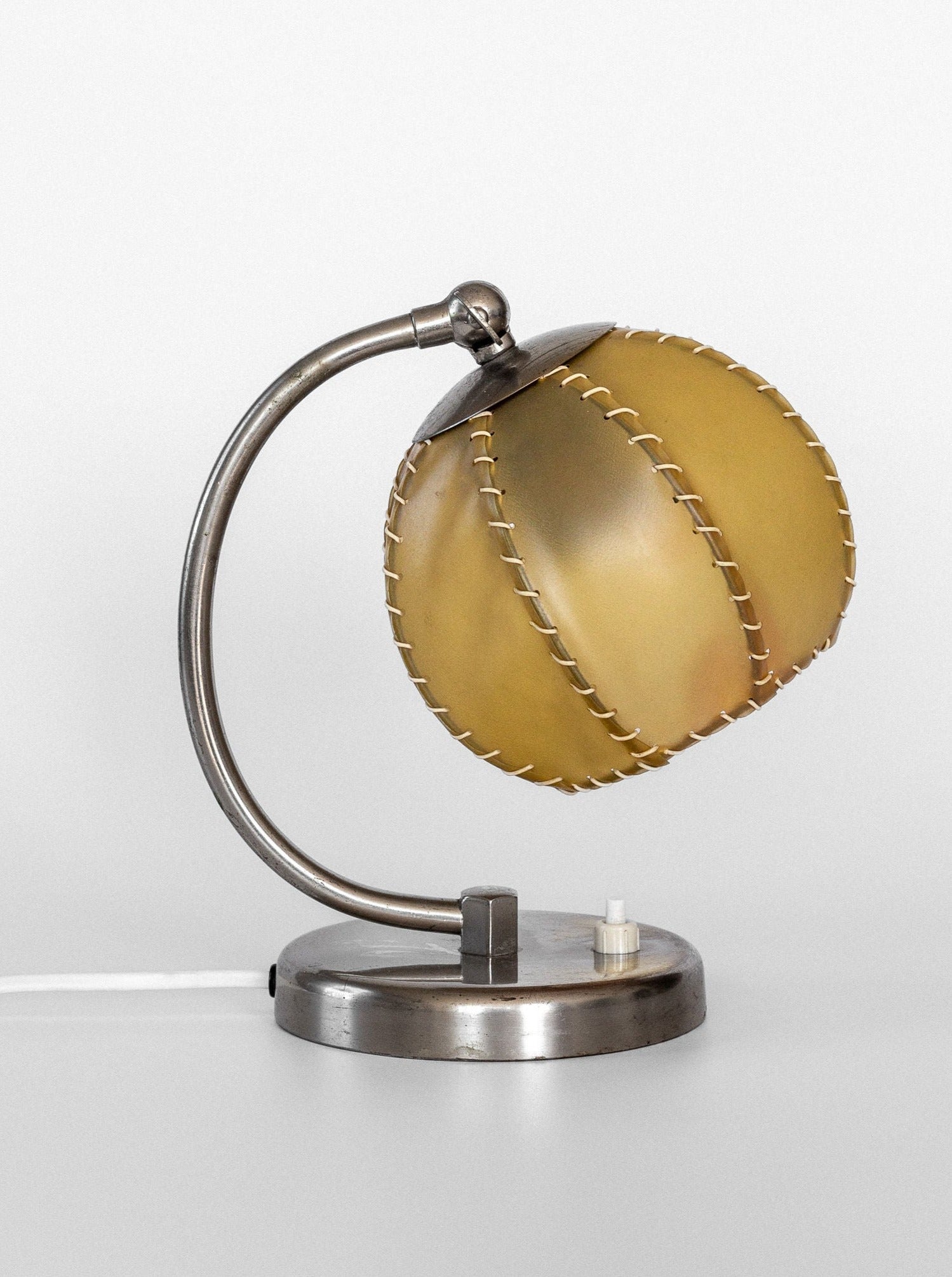 This collectible vintage table lamp, the 1930s Art Deco Metal Lamp by Spigel, features a curved metal stand and a round, stitched yellow lampshade with a unique design resembling hand-stitched leather. It is set on a simple chrome body base with a switch, against a plain white background.