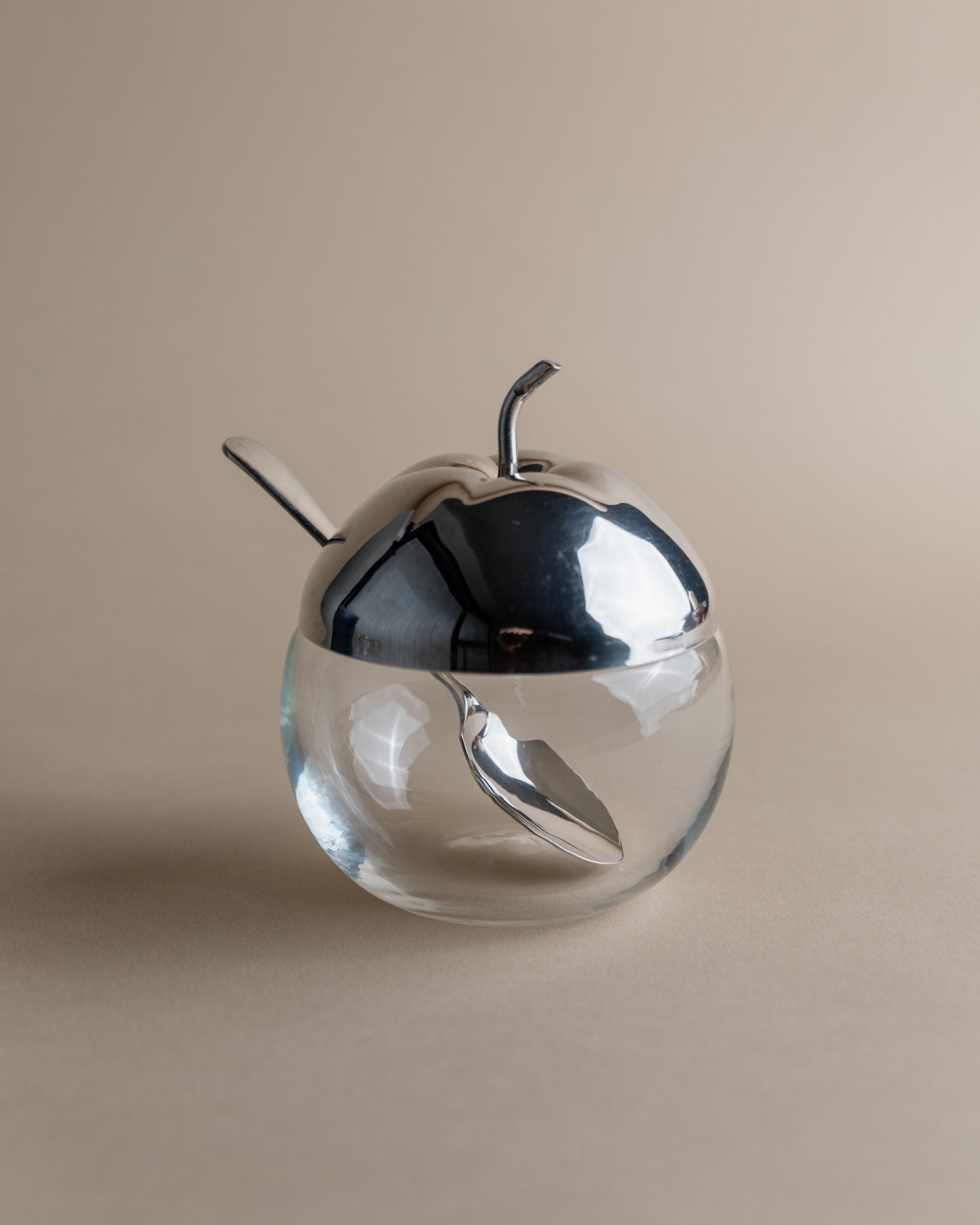 The Silver & Glass Apple Jar by Bottega Jacobs doubles as a sugar bowl, featuring an apple-shaped, silver-plated lid and a matching spoon inside. Its minimalist elegance is enhanced by the soft beige background.