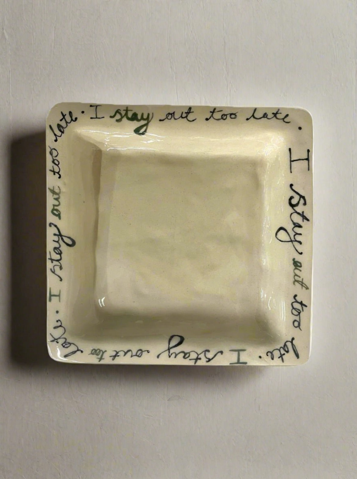 The "I Stay Out Too Late Dish" by L&Clay Ceramics is a square, beige ceramic bowl featuring a glossy finish. It showcases a handwritten inscription along the rim that repeats the phrase "I stay out too late" in green and black lettering. With its unadorned center, this custom-made piece adds a unique touch to any collection.
