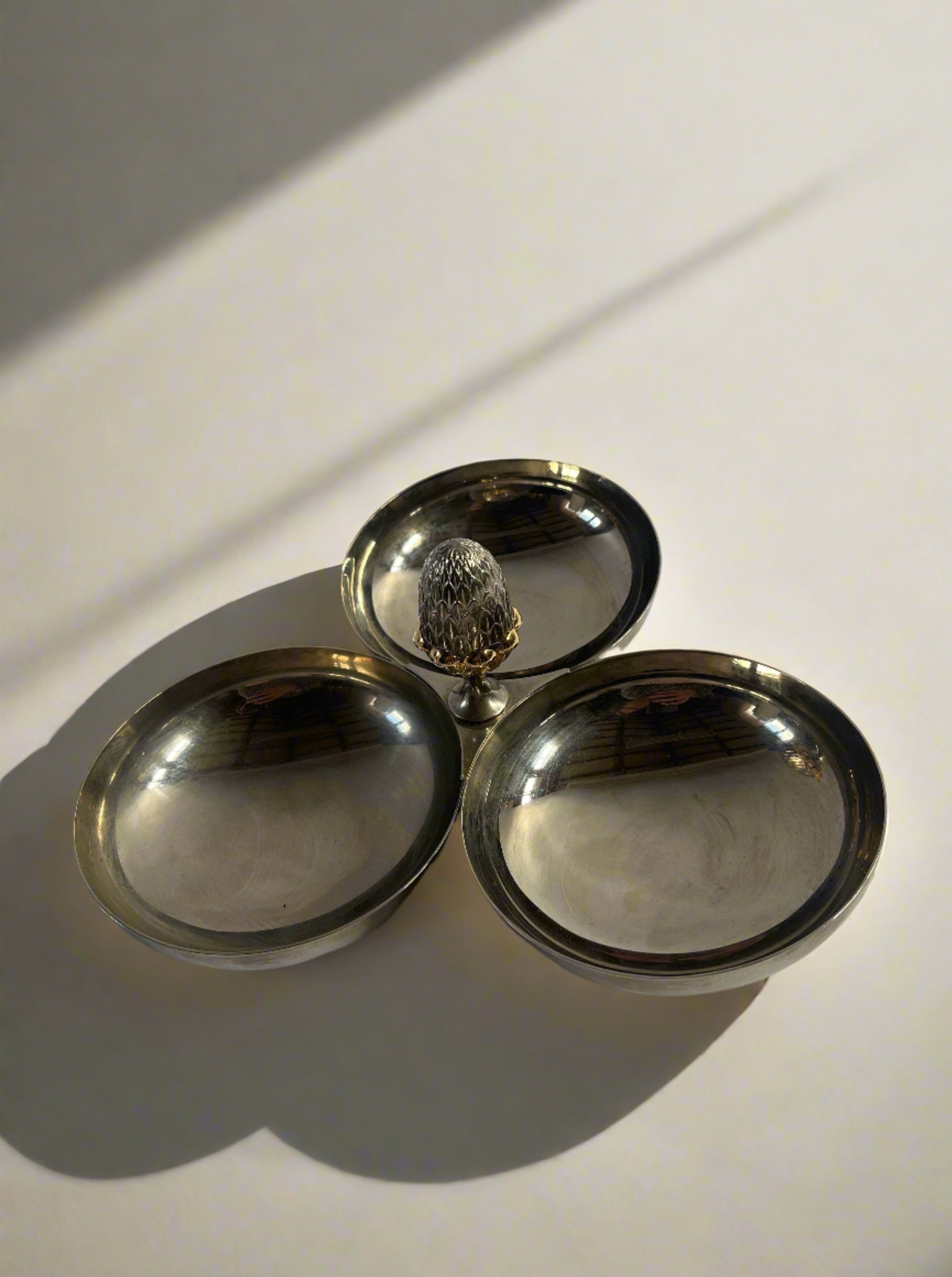 A Maud Vaughan Vintage French Nickel and Brass Serving Dish with three round, polished compartments connected at the center. The nickel finish is reflective and elegant, and the center features a small, decorative brass acorn handle. The background is plain and white.