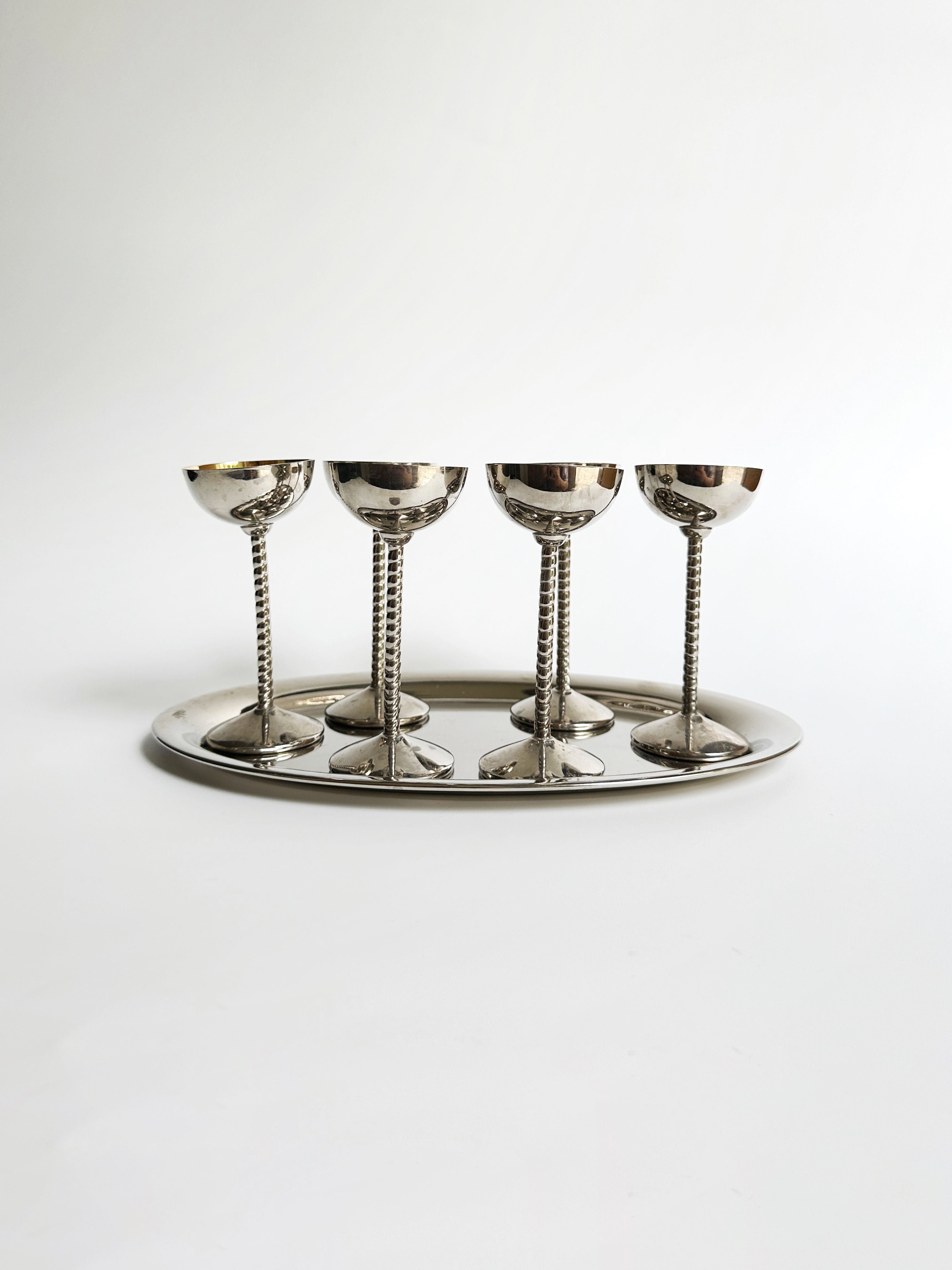 The Art Nouveau liqueur goblet set by Dodo Vintage features a quartet of silver goblets with intricate stems, evoking the elegance of the Art Nouveau era. They are beautifully arranged on an oval silver tray. Against a plain white backdrop, the gleaming reflective surfaces of both the goblets and tray are showcased, creating an aesthetic that is both minimalist and refined.