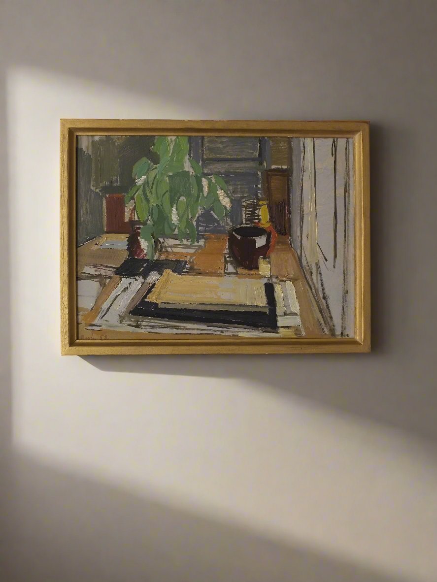 An Interior Oil Painting by Swedish artist Sven Olof Rosen, dating back to 1958 and available through Veter Vintage, hangs in a wooden frame on a beige wall. The oil on board artwork depicts an abstract still life with various objects, including a green plant, scattered on a table covered with patterned cloths. Sunlight casts a shadow on the wall adjacent to the painting.