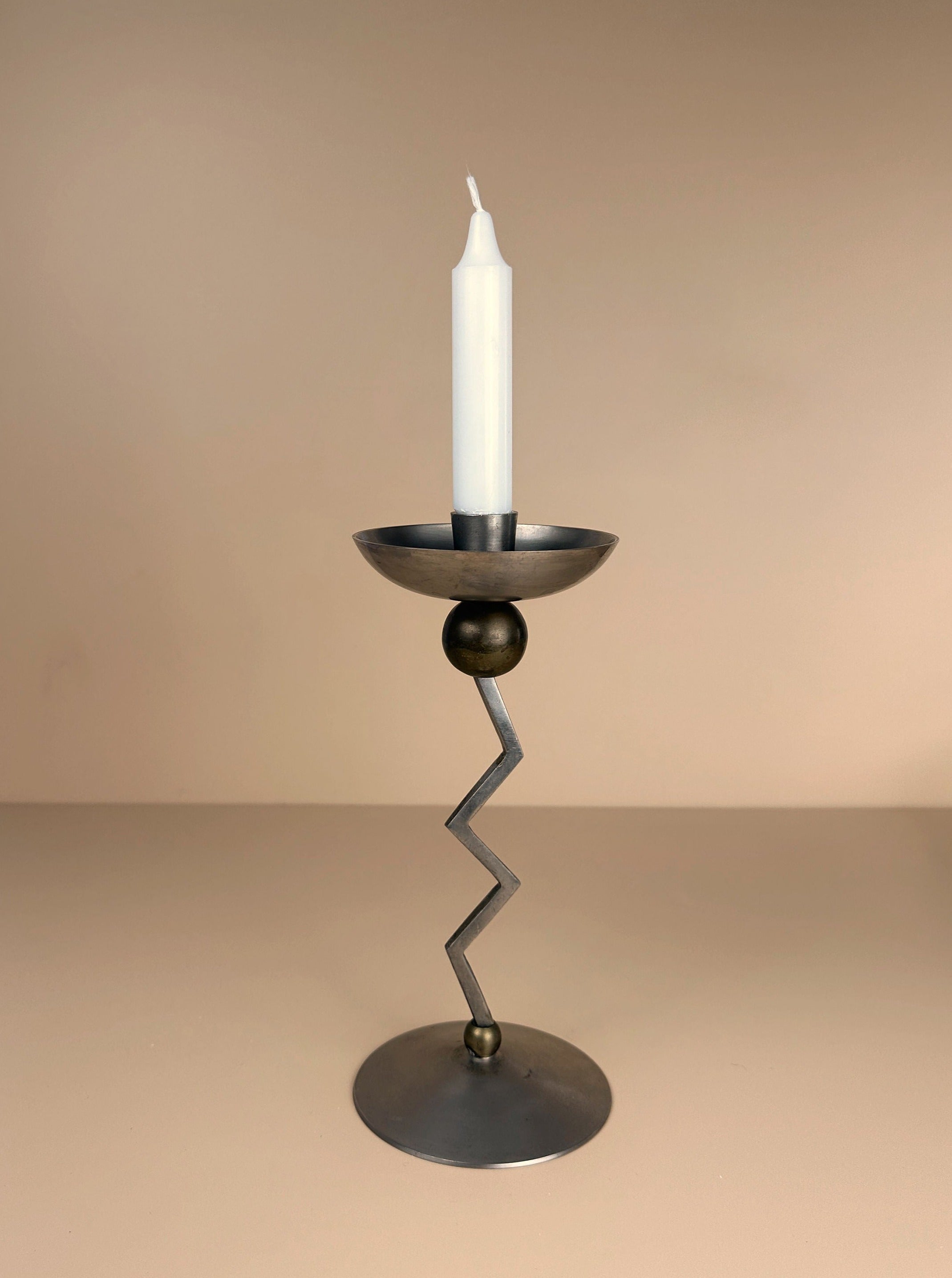 A sleek, modern Postmodern Zig Zag Candle Holder by Dodo Vintage, featuring a silver-colored metal stem and circular base, is centered in the image. It holds a tall, tapered white candle against a soft, warm beige background, evoking Memphis-style design elements.