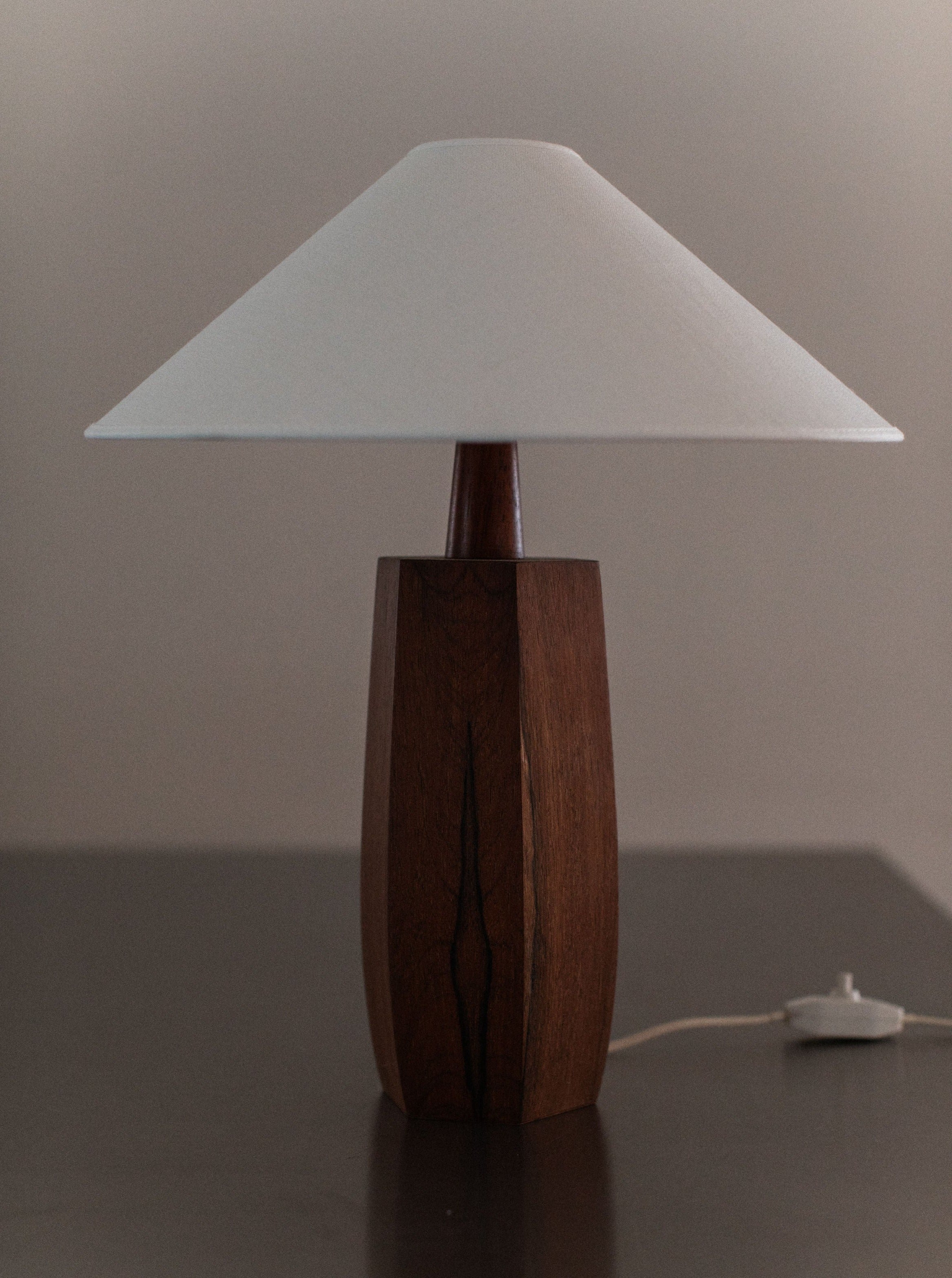 A 1940's Wooden lamp from Out For Lunch, showcasing mid-century design with a white, conical shade, stands on a dark surface. The lamp's cord and switch are visible at the base, extending towards the right. The background is neutral, highlighting the lamp's simple and elegant craftsmanship.
