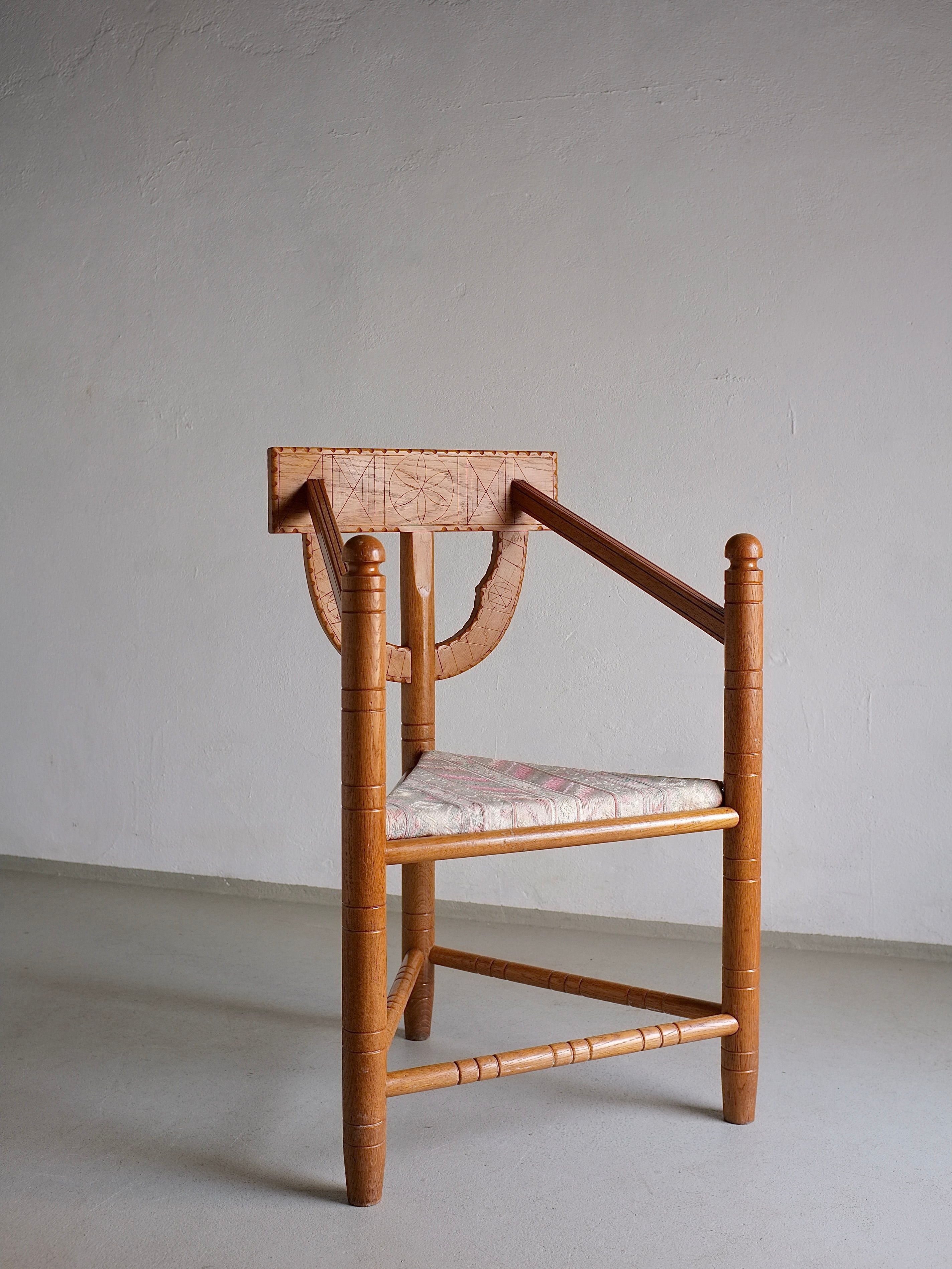 The Rustic Carved Oak Corner Chair from Veter Vintage features a unique, vintage design with a geometric backrest and angled armrests. This wooden chair has a patterned cushioned seat and spherical accents on the legs, reminiscent of Sweden's 1950s design period. It is pictured standing against an off-white wall with a gray floor.