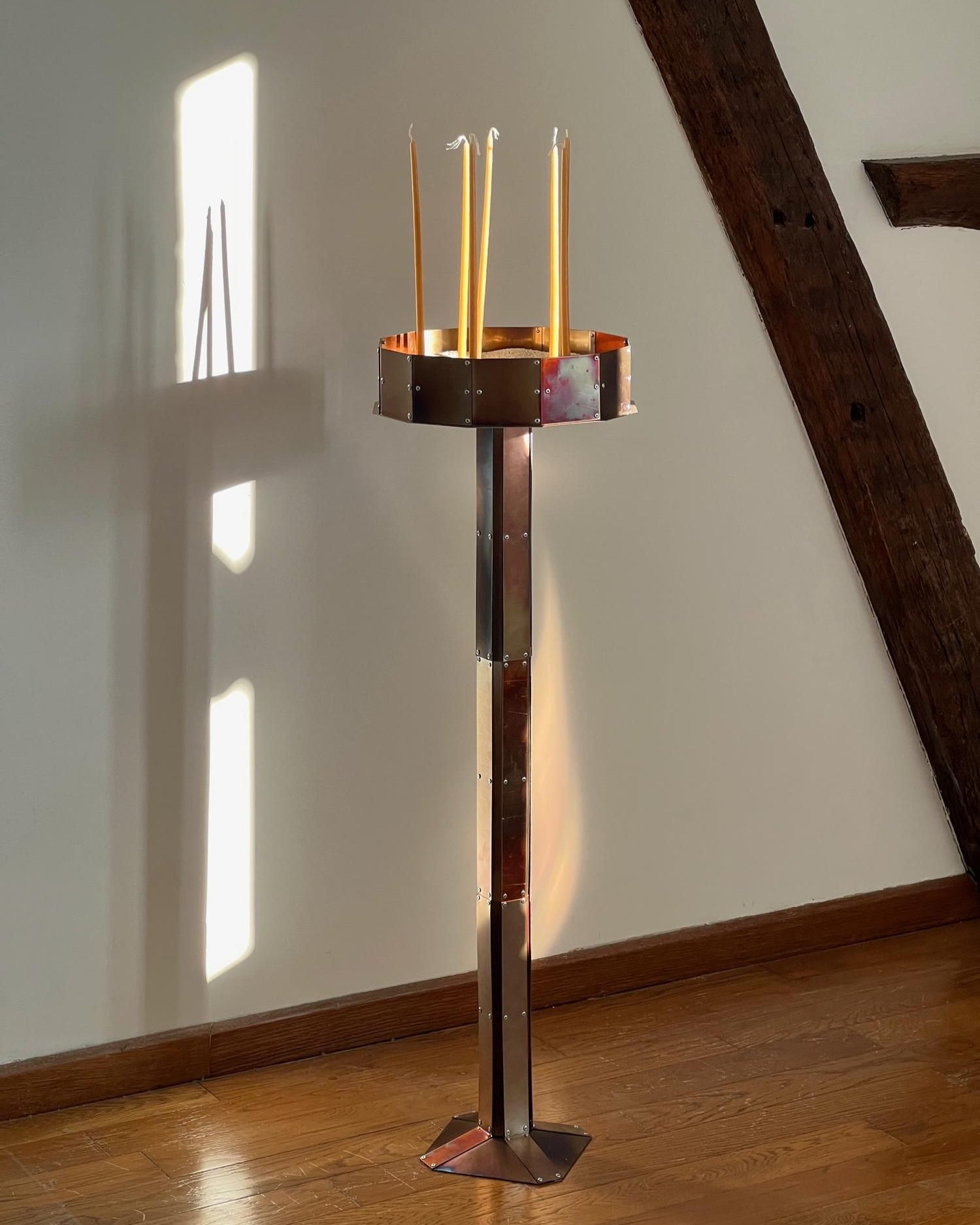 The Manouali candleholder by Theo Galliakis, a tall metallic Orthodox Christian piece, stands on a wooden floor. Four unlit taper candles cast soft shadows as sunlight spills across a white wall with wooden beams.
