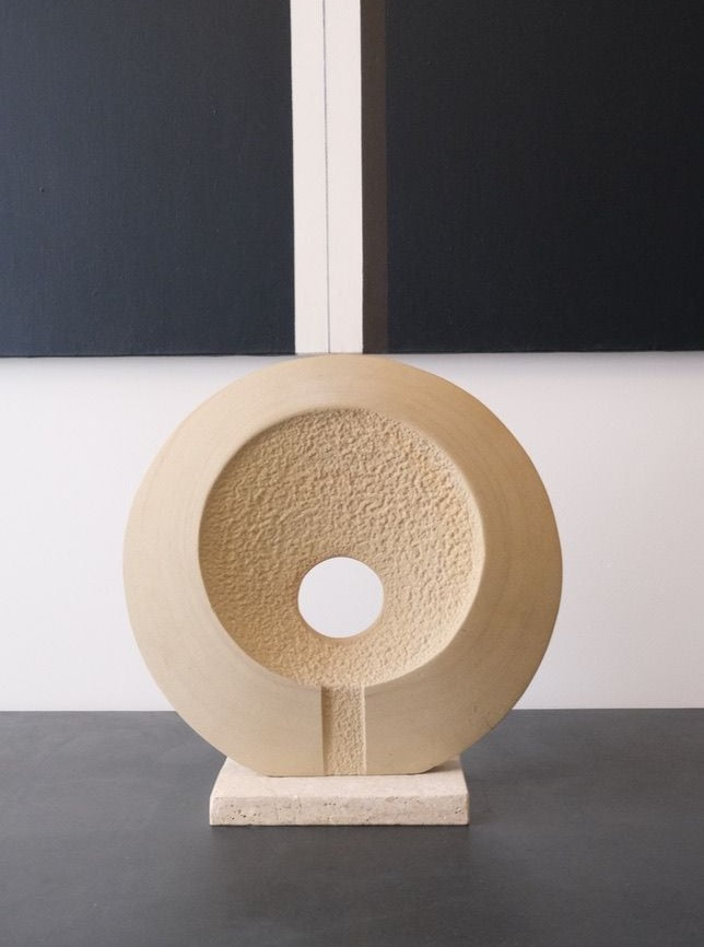 A mid-20th century, circular Abstract Limestone Sculpture by LA NOUVELLE GALERIE with a rough-textured inner surface and a hole in the center sits on a travertine base. It is positioned on a dark surface with a minimalistic background of grey and black panels.