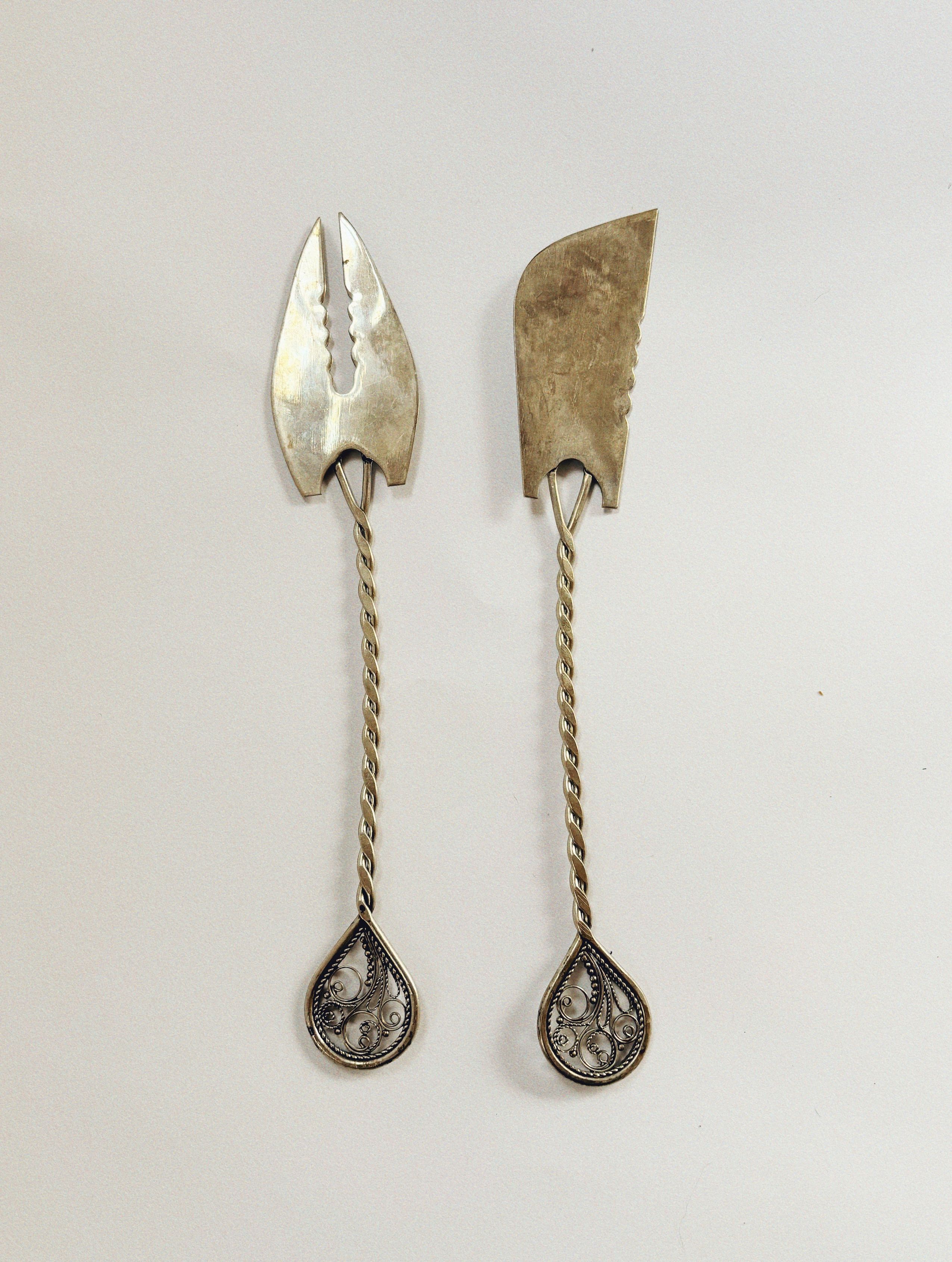 Presenting the Silver Plated Serving Cutlery by Huerto: two exquisite utensils characterized by intricately detailed handles against a plain backdrop. The handles feature beautifully twisted stems and teardrop-shaped ends embellished with delicate filigree designs. Ideal for enhancing any table setting, these pieces also have uniquely shaped heads with serrated edges.