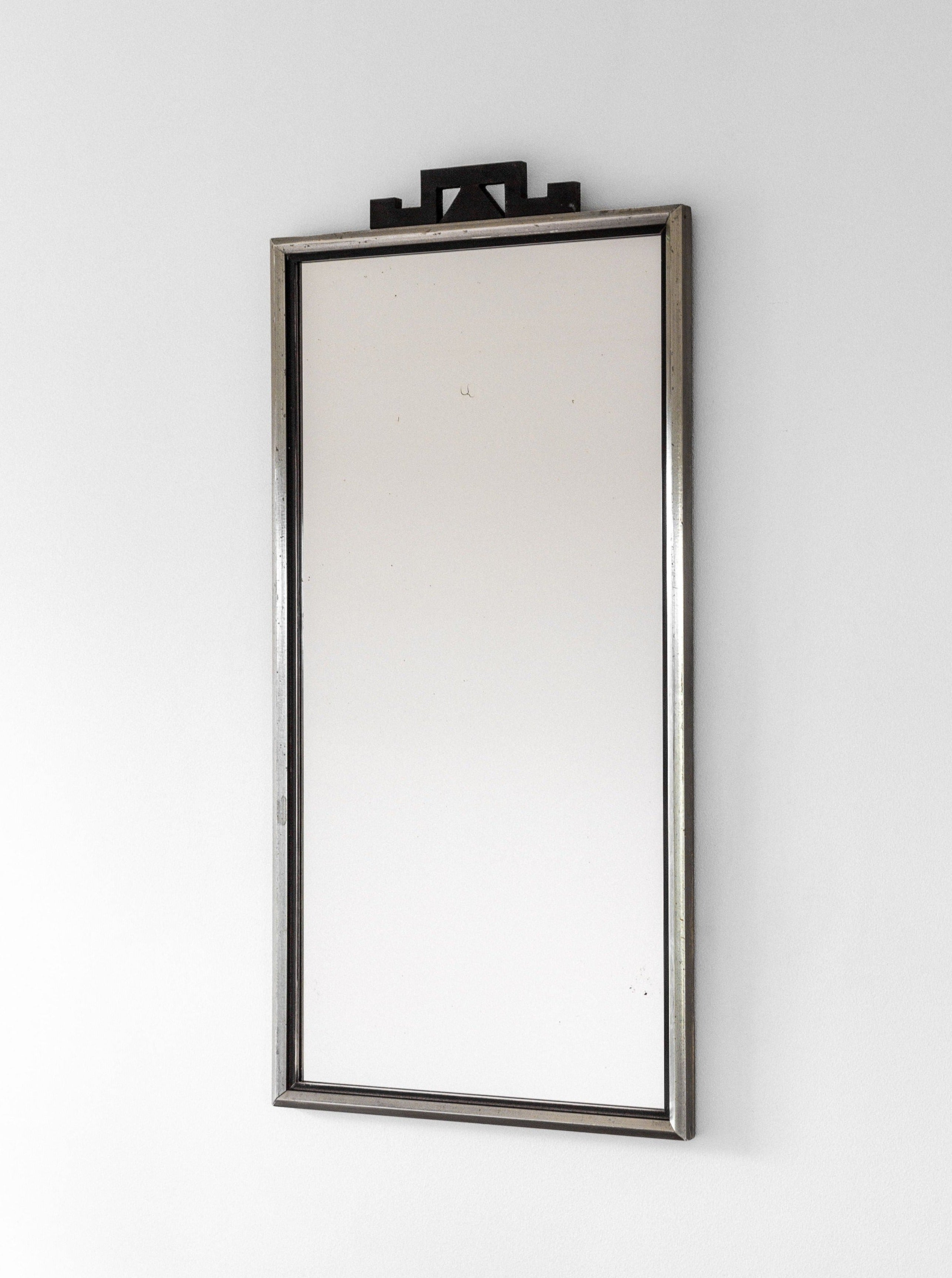 A 1930s Swedish Art Deco mirror by Spigel features a thin, metallic frame and an ornate, black decorative piece on top. The mirror is mounted on a plain white wall and reflects a neutral, empty space.