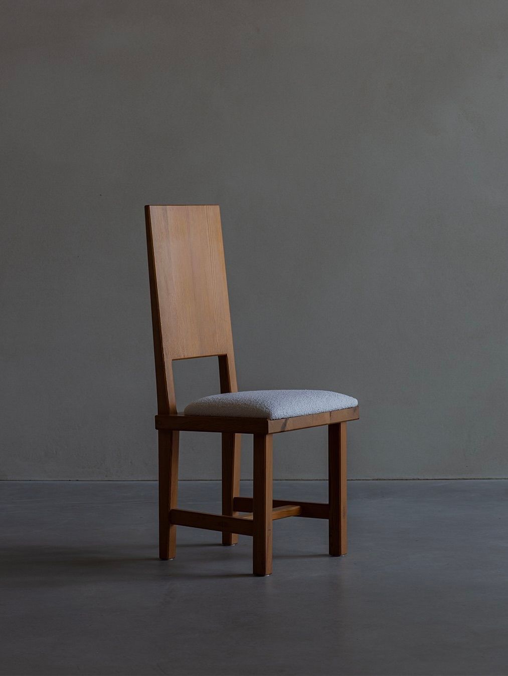 A Göran Malmvall - Model 553 chair by Bicci de' Medici, featuring a wooden frame, tall rectangular backrest with a square cut-out, and light gray padded seat, stands against a dark gray backdrop, capturing the essence of Swedish minimalist design.