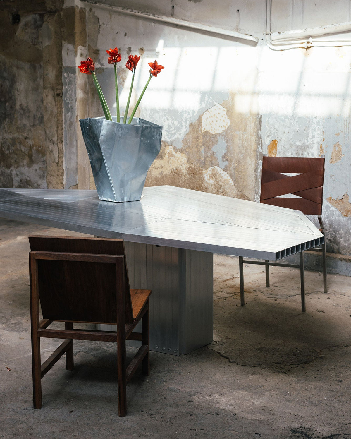 An Aluminum Dining Table by Studio HAOS stands in a weathered industrial room. A geometric vase with red flowers sits on top, flanked by two mismatched chairs, reflecting the brand’s signature contemporary design.