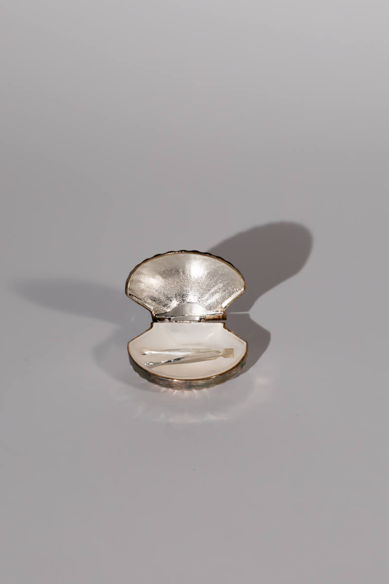 The Vintage Shell Pill Box by Break The Mould Magasin is a small, seashell-shaped compact crafted from alpaca metal, featuring a mirrored interior. Its metallic exterior casts a shadow on the light gray background. This vintage pill box gleams within, making it a collector's treasure.