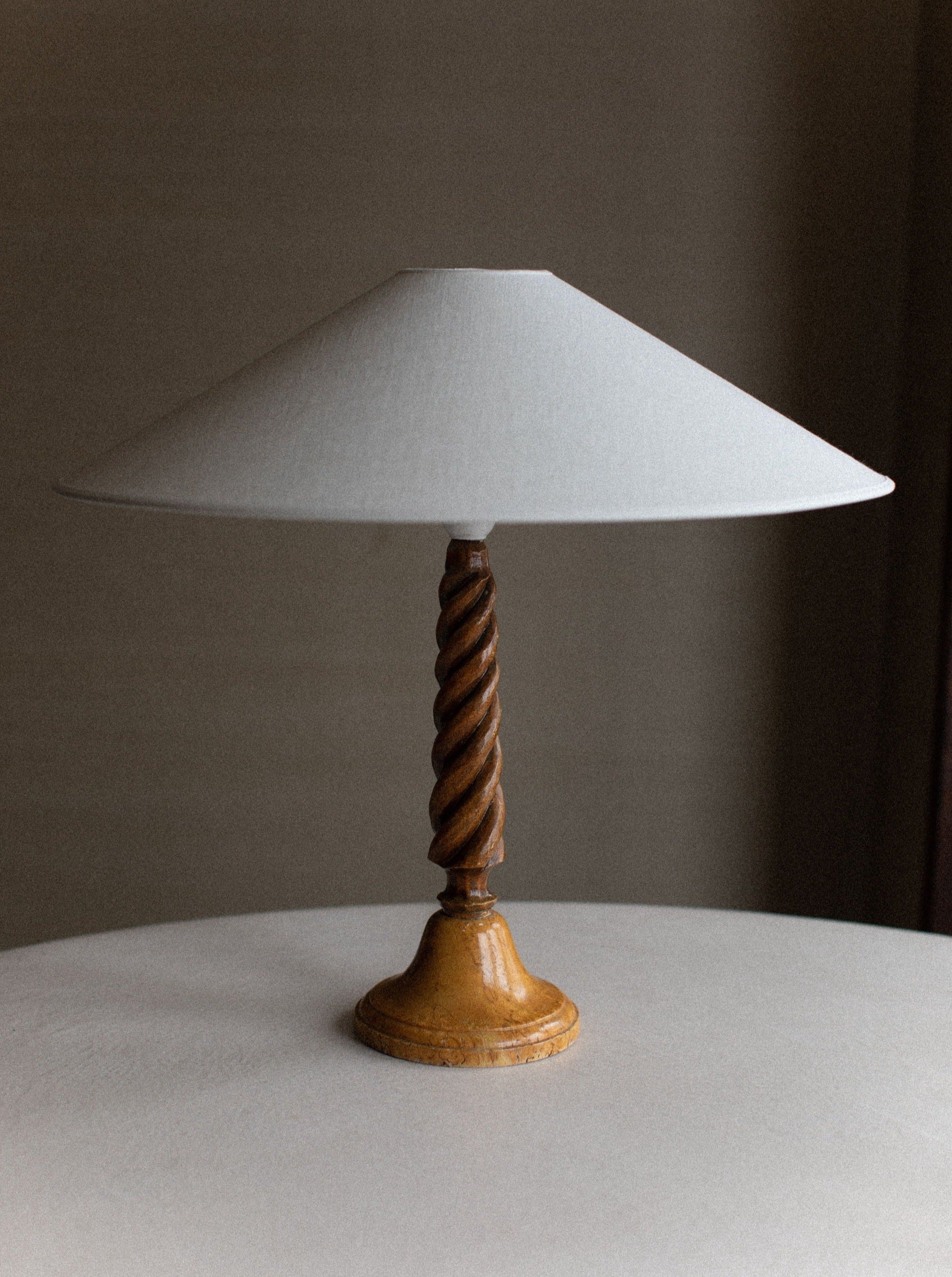 A vintage Twisted Wooden Lamp with a white, cone-shaped lampshade from Out For Lunch sits on a round, light-colored surface. The lamp features a wooden base and a spiral-carved stem, showcasing artisanal craftsmanship and the natural wood grain. The background is neutral, with soft, diffused lighting.