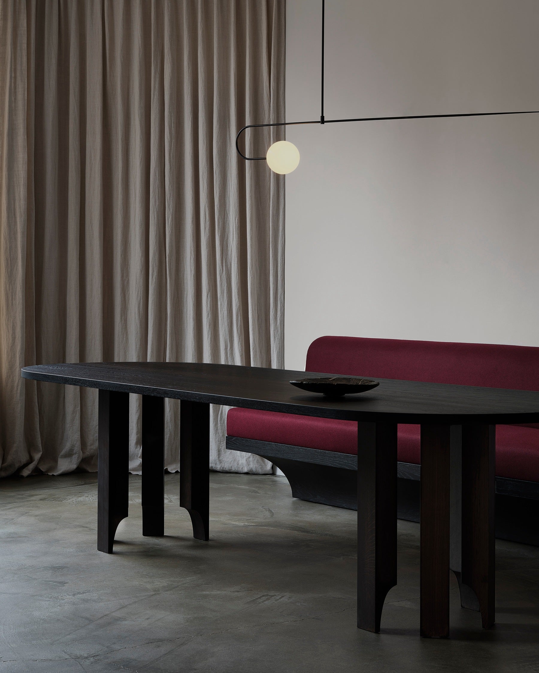 A minimalist dining room with a customizable design features the Mokko Acer Capsule Table, a dark red cushioned bench, sheer beige curtains, a modern black pendant light with a round bulb, and a bowl on the table atop a gray concrete floor.