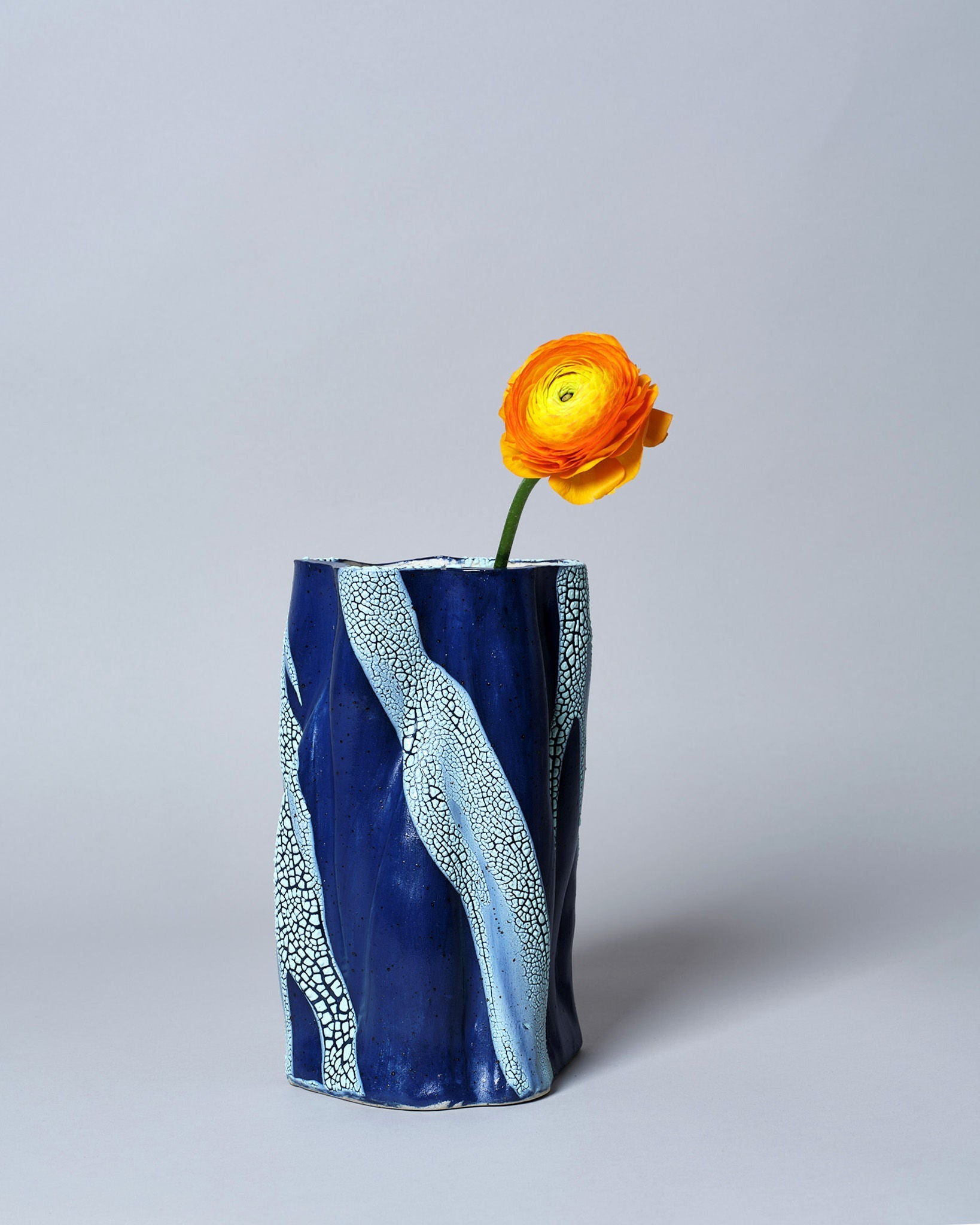 A single yellow and orange ranunculus in the tall, handmade "Swirly Vase Blue and Light Blue" by Astrid Öhman, featuring blue and light blue patterns, set against a plain light gray background.