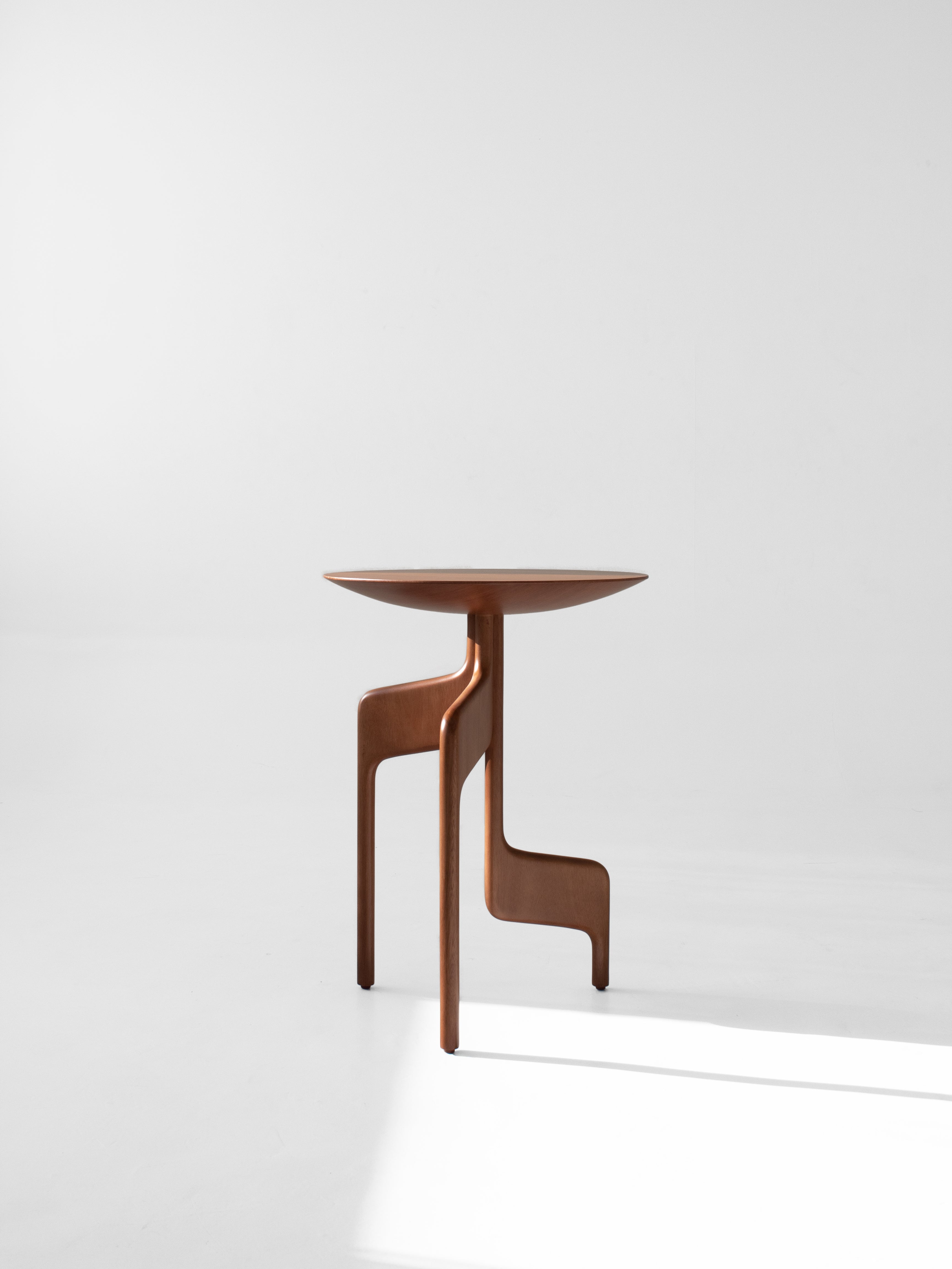 The "Vereda" Side Table by Luciano Costa Estúdio is a modern, minimalist wooden piece with a round top and three asymmetrical legs, showcased against a plain white backdrop.