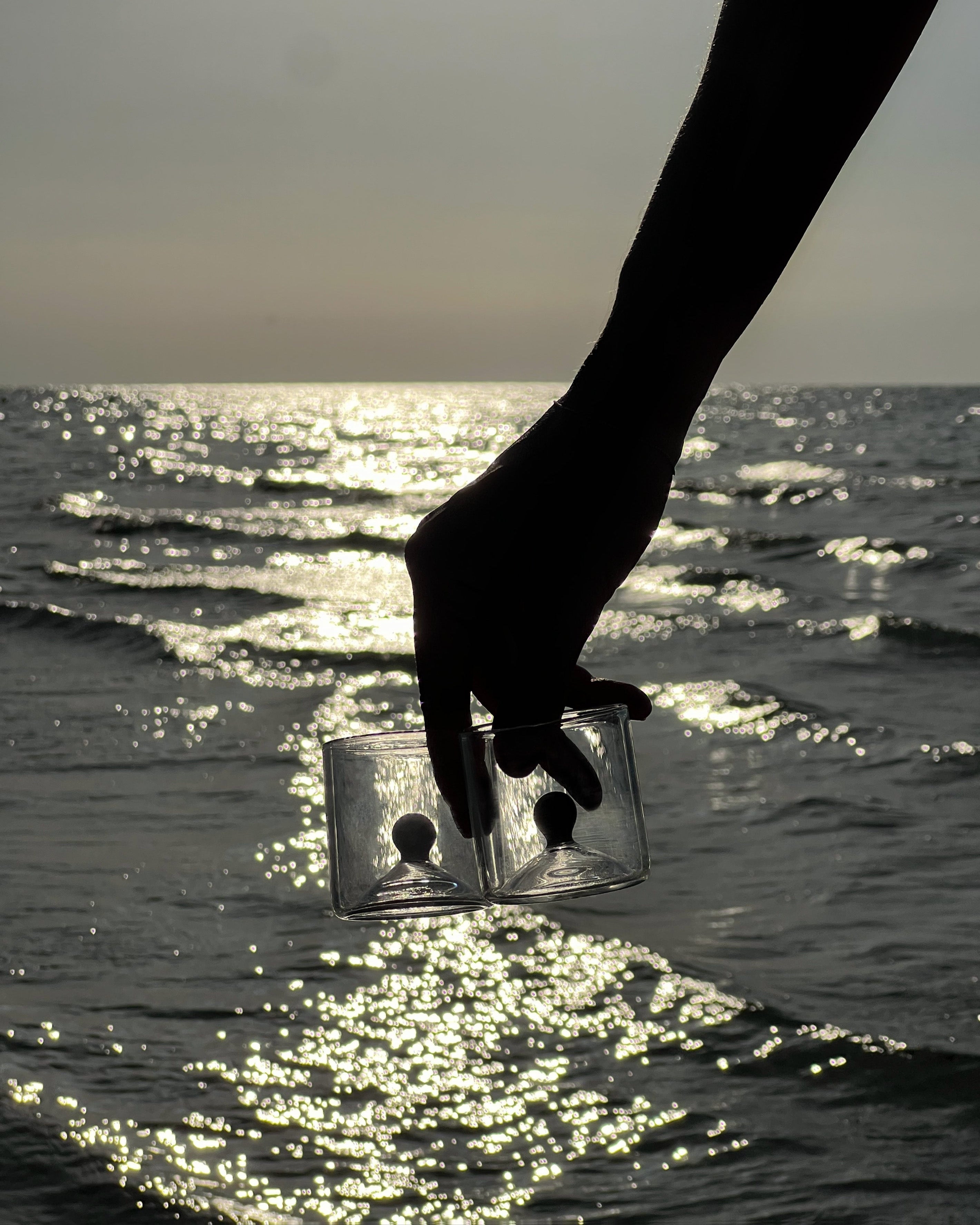 Hand holding green glass shot duo set against sparkling ocean water