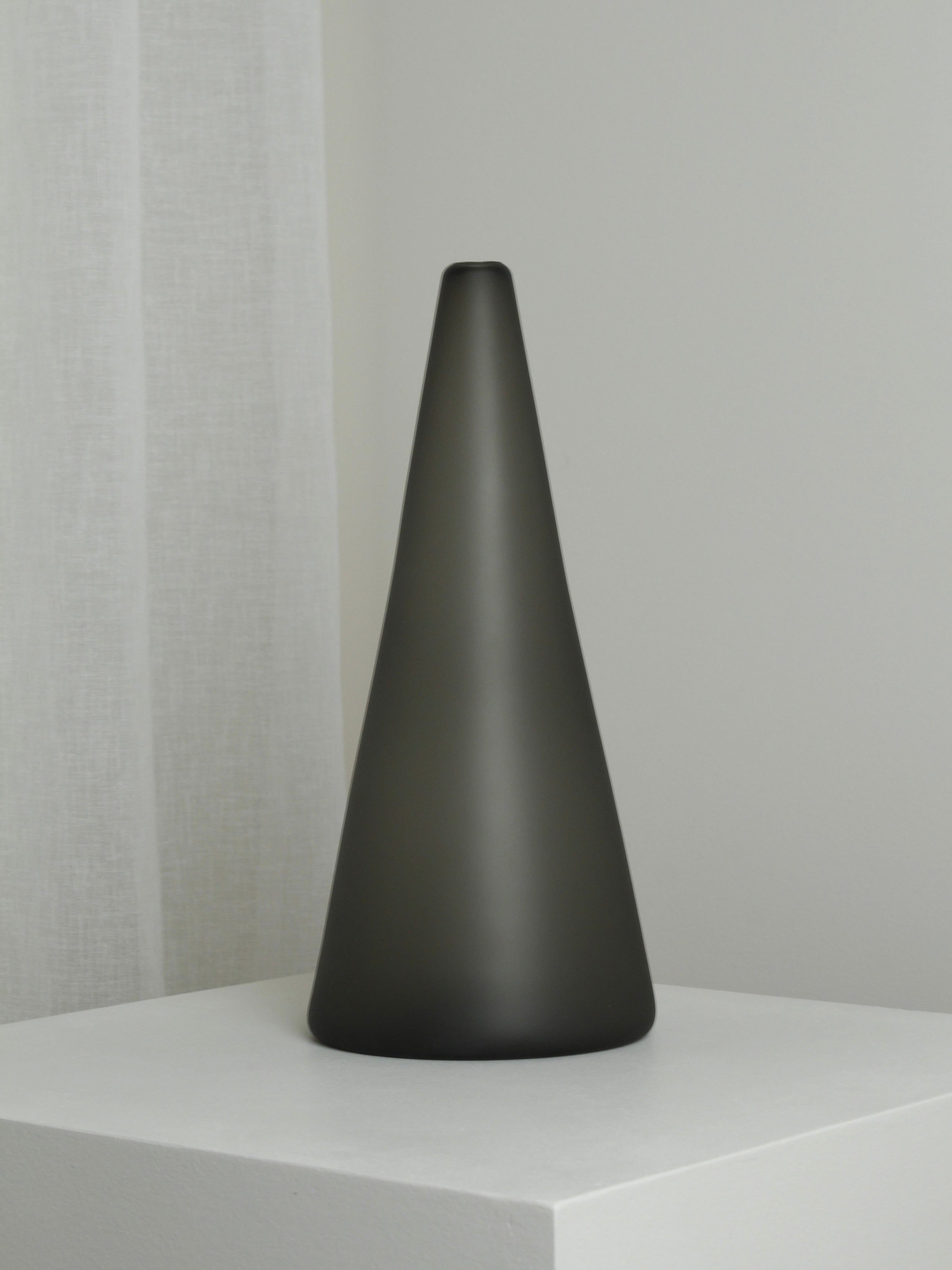 The XLarge Vase in Smoke by Verre d'Onge, featuring a cone shape and matte black finish, sits on a white pedestal in a minimalist room with white walls and a sheer curtain.