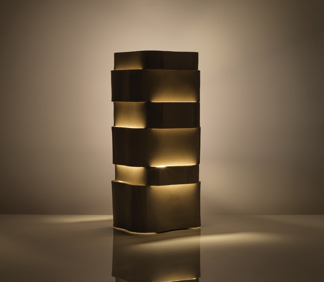 The Too 60-2 Table Lamp by Katia Langeard features a tall, rectangular ceramic design with stacked, uneven segments, emitting a warm, soft light ideal for bedside use and creating gentle shadows on nearby surfaces and walls.