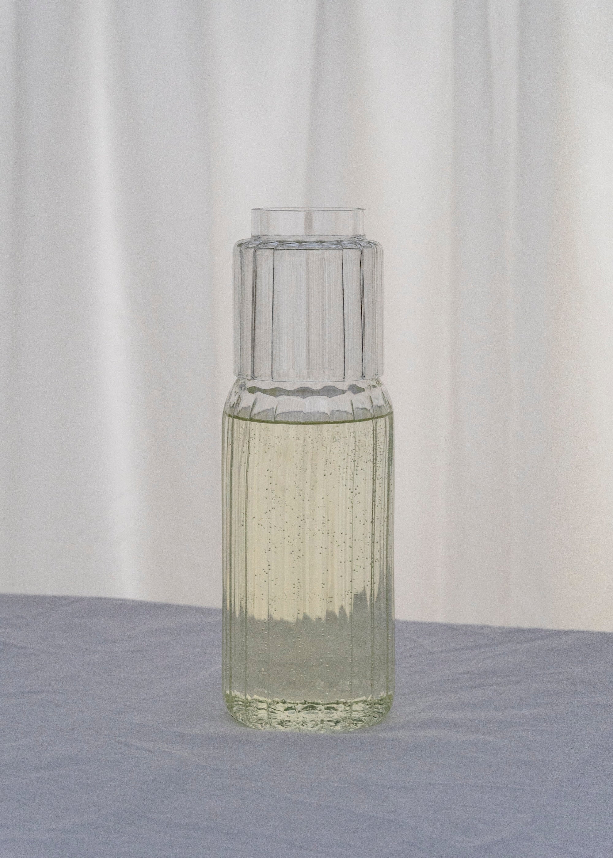 The Pillar — Carafe Set by Agustina Bottoni, handcrafted in Italy from borosilicate glass, features vertical ridges and includes a matching glass lid. The clear set is shown with a bubbly drink atop blue-gray fabric and a white curtain backdrop.