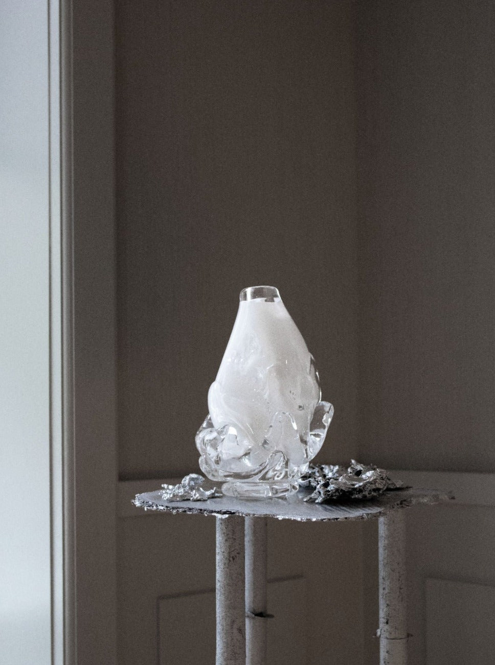 The Szkło Studio Opal Vase - Small, featuring an abstract organic glass design, sits on a rustic white table next to a textured silver object, bathed in soft natural light against a neutral-toned wall.