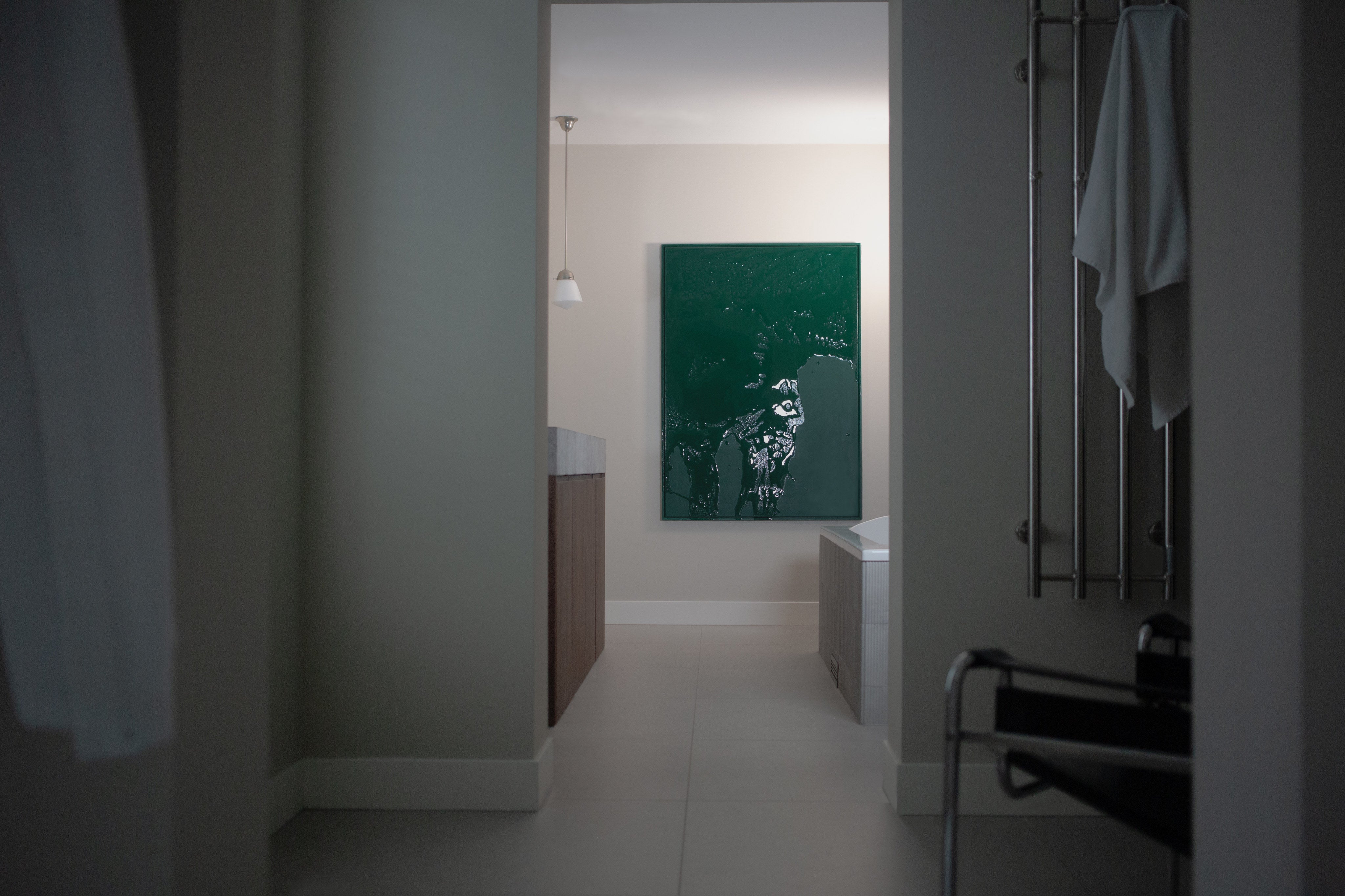 A modern, minimal bathroom with neutral tones features STUDIOPOLS' Green Flux – Uncategorized sculptural wall art in dark green on the far wall, a freestanding tub, a towel rack, and soft natural light streaming from above.
