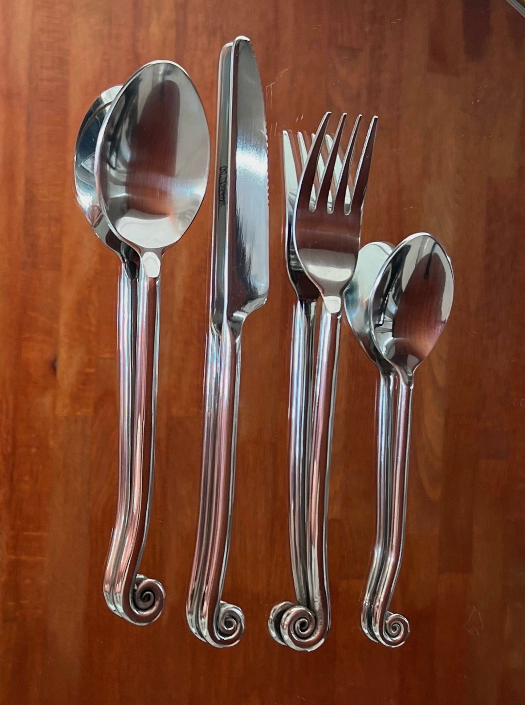 A set of sophisticated cutlery featuring a spoon, knife, fork, and a smaller spoon. Each piece in this Manushi Set of Vintage Swirl Cutlery has a distinctive curved handle design with decorative spiral ends, laid out neatly on a wooden surface. The utensils reflect light, giving a polished appearance.