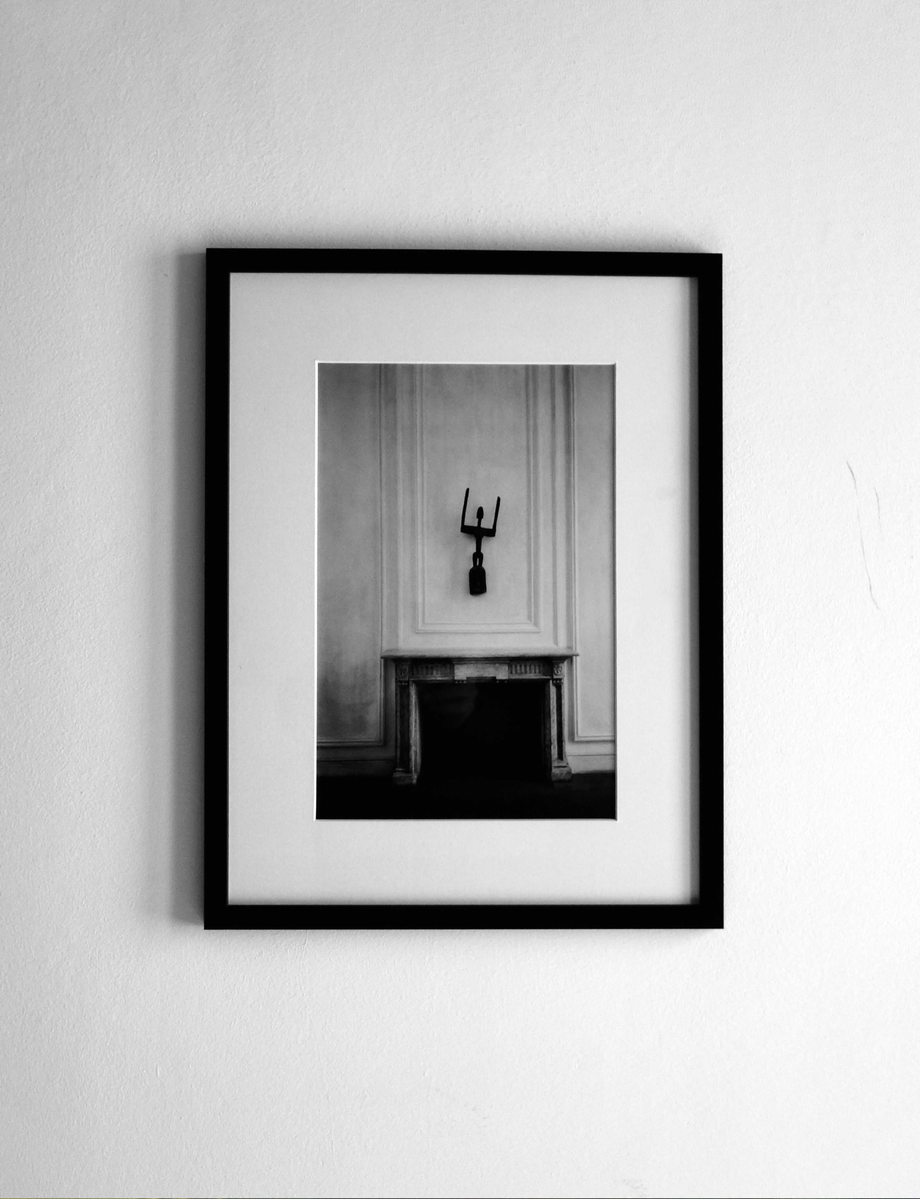 COSEINCORSO's "Memory #03" contemporary black-and-white photographic print, in an aluminium frame, features a decorative mask above a mantelpiece, displayed on a white wall in an elegant, minimalist setting.