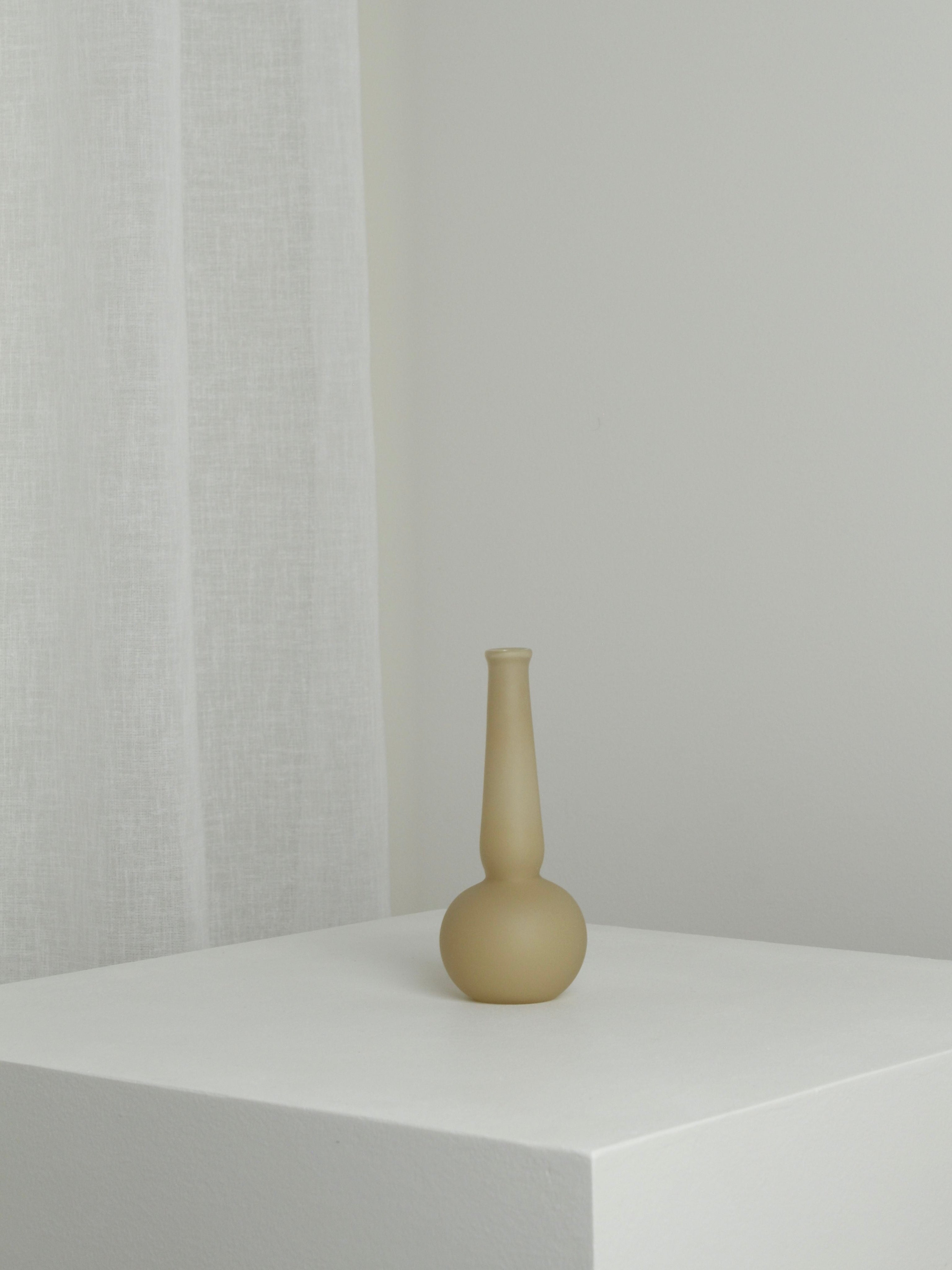 The Verre d'Onge Small Vase in Caramel, featuring a matte finish and long neck, is displayed on a white pedestal against a light wall and sheer white curtain, creating a minimalist, uncluttered setting.