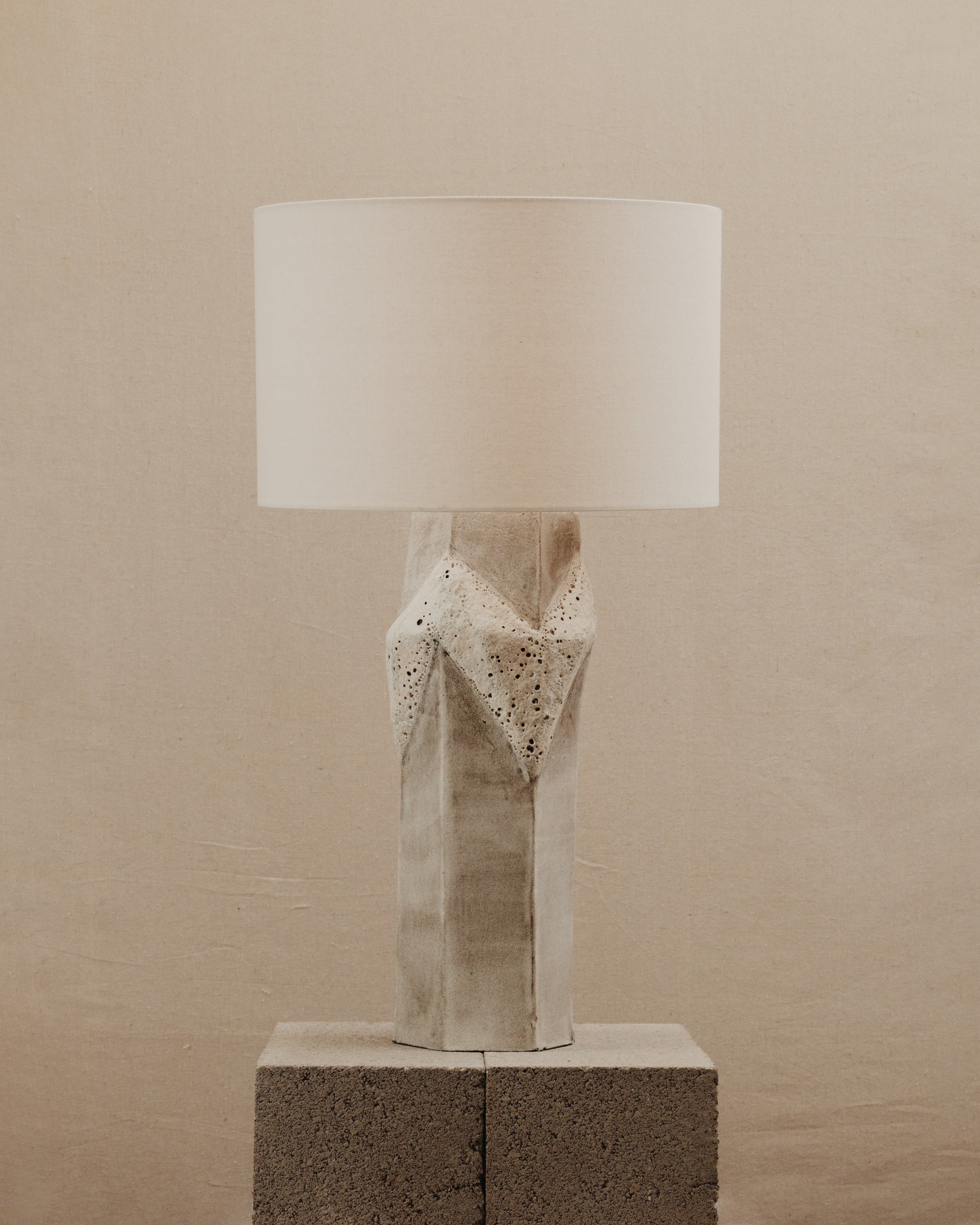 The JS/WRKSHP Warp Table Lamp, featuring a sculpted ceramic base and white fabric drum shade, is displayed on two stacked cinder blocks against a beige background.