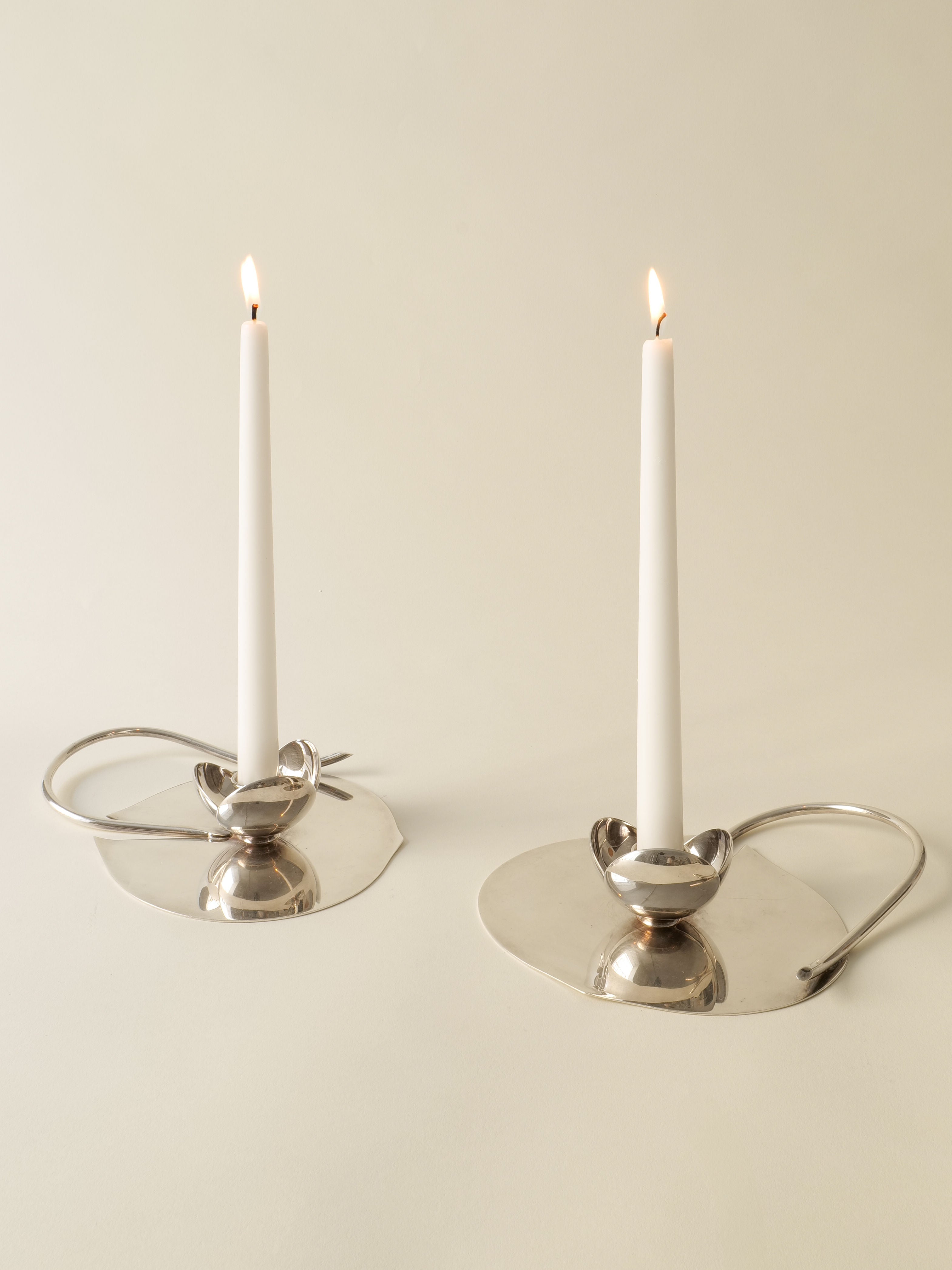 Two lit white candles in sleek, modern Candle Holder Lino Sabattini 1970s by Collection apart, featuring silver-plated wide flat bases and curved handles, placed on a neutral surface against a light beige background.