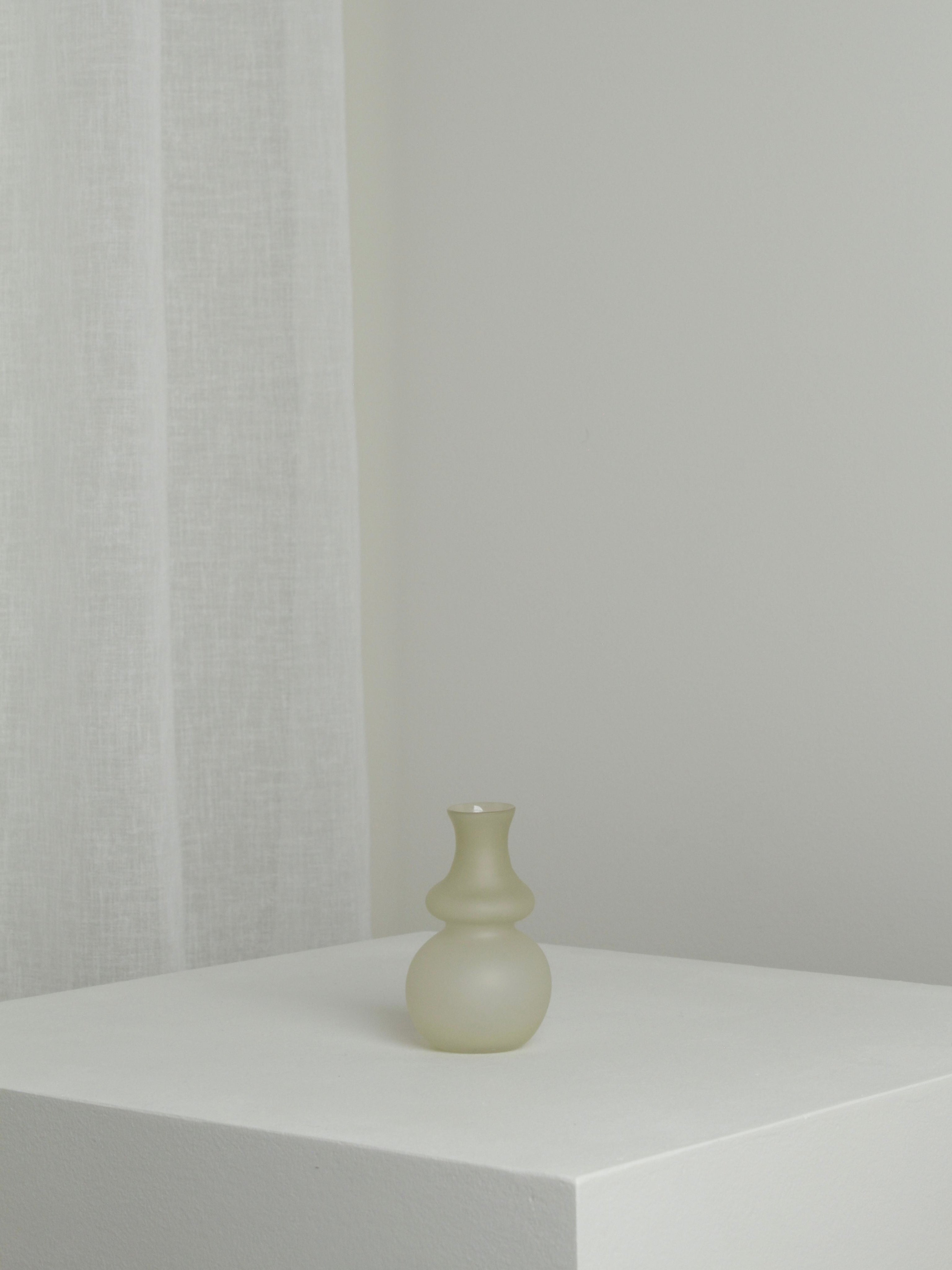 The Small Vase in Stone by Verre d'Onge, with its frosted glass and rounded base, sits on a white pedestal in a bright, minimal room. A sheer curtain in the background highlights this delicate vase's elegant beauty.