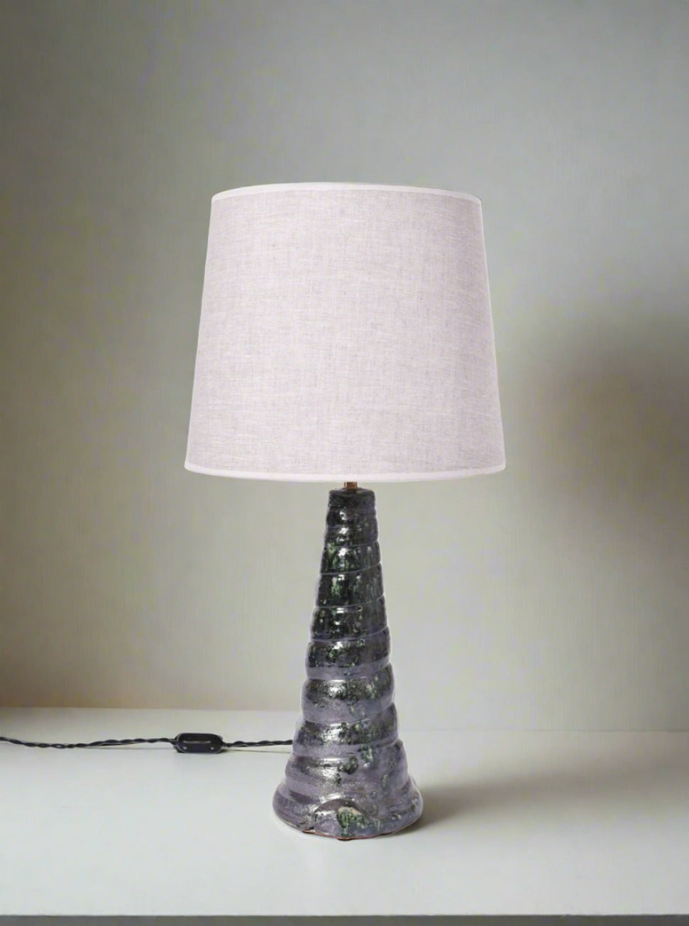 The "Teotihuacan" Green Ceramic Table Lamp, Barracuda Edition by Barracuda Interiors, features a textured spiral ceramic base inspired by Teotihuacan artifacts and a plain fabric shade. It sits on a white surface with its cord and switch visible.