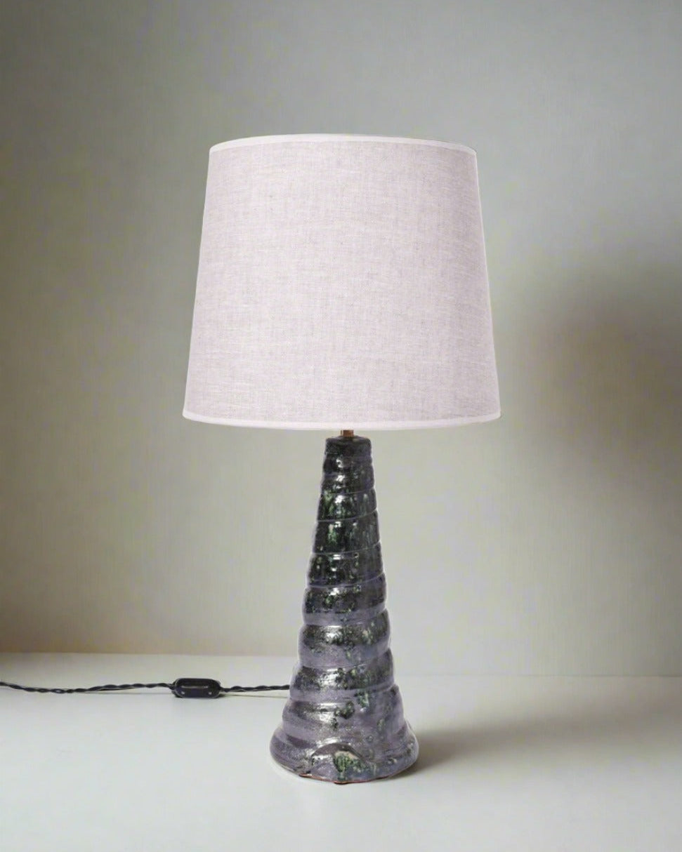 The "Teotihuacan" Green Ceramic Table Lamp, Barracuda Edition by Barracuda Interiors, features a textured spiral ceramic base inspired by Teotihuacan artifacts and a plain fabric shade. It sits on a white surface with its cord and switch visible.