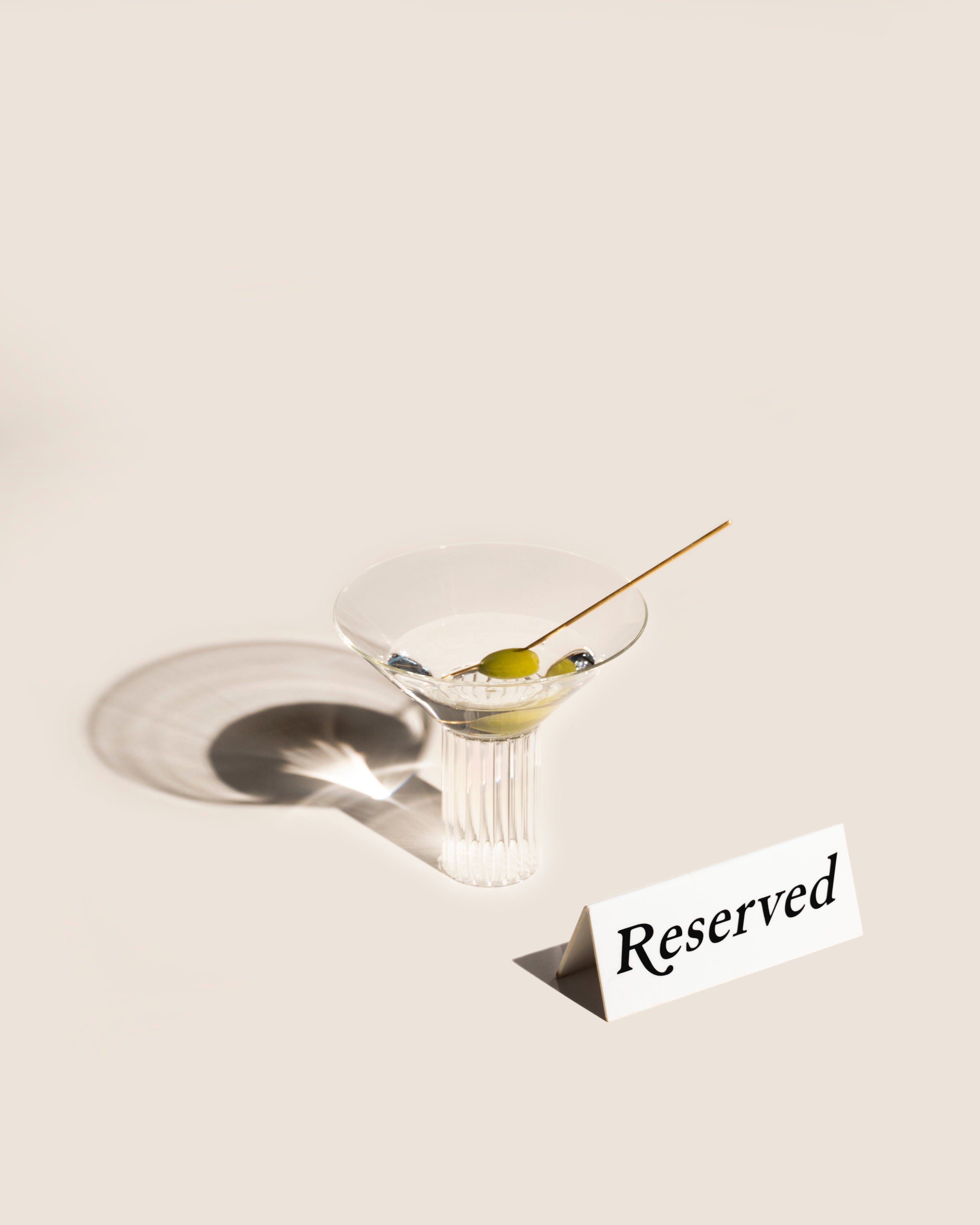 The Calici Milanesi Cocktail Glass by Agustina Bottoni, handmade in Italy from borosilicate glass, holds an olive on a garnish stick beside a white "Reserved" card—all set against a neutral background with a distinct shadow.