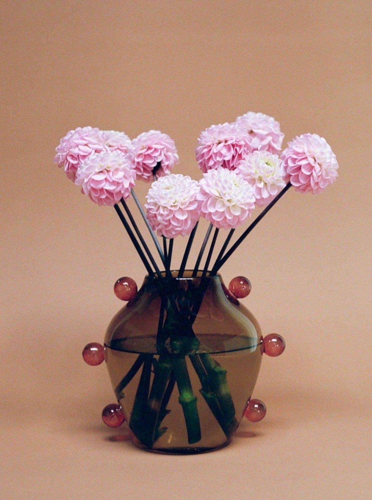 A decorative La Chaleur Vase from Sophie Lou Jacobsen adorned with abstract patterns, containing pink chrysanthemums and ornamented with spherical glass accents on thin stems, set against a pastel peach background.