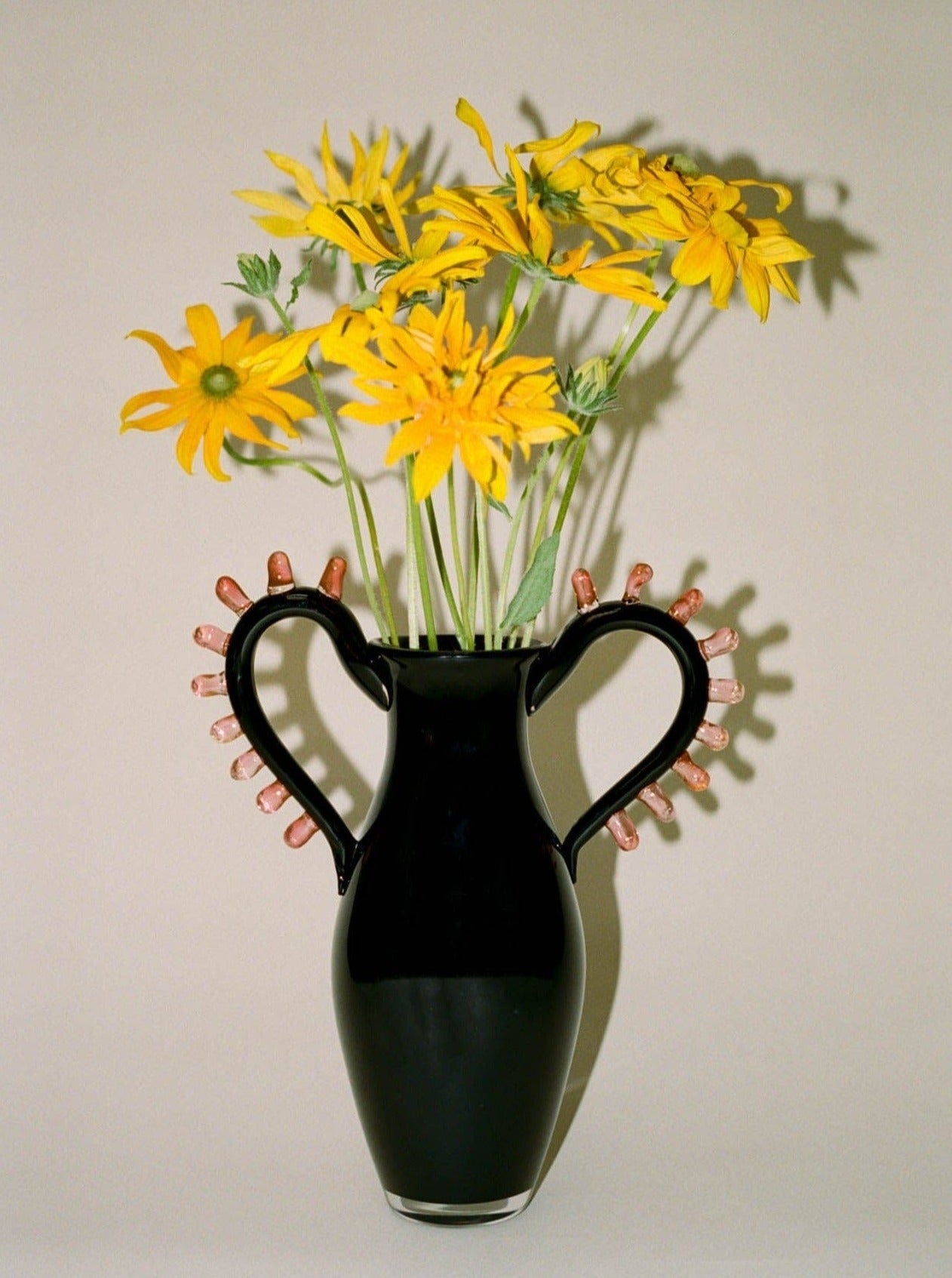 The L'Aurore Vase by Sophie Lou Jacobsen, featuring sculptural handles and decorated with small objects, holds yellow flowers with green stems against a simple backdrop.