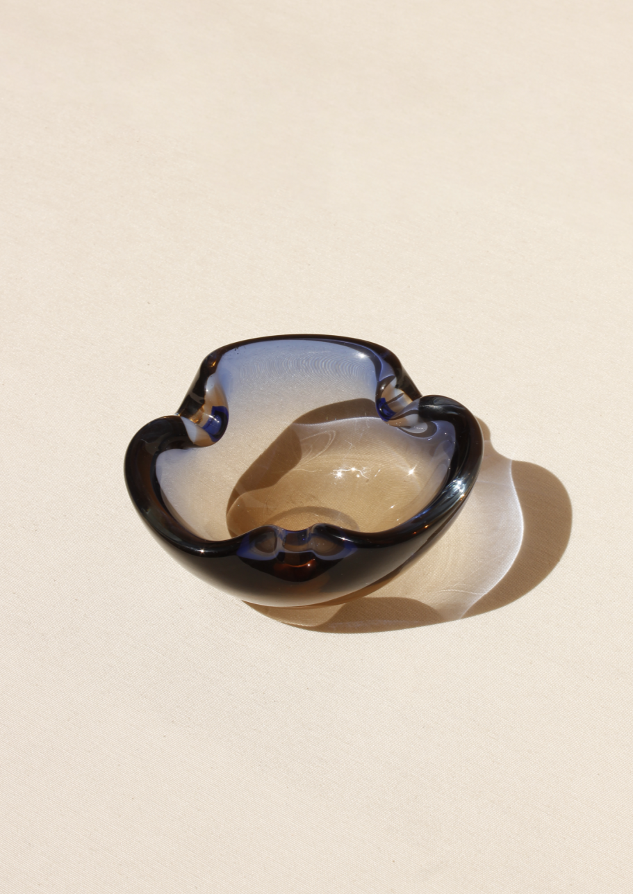 The Two-Colour Murano Bowl from Boga Avante Shop is a small, elegantly designed black and clear glass piece with smooth curves. Crafted for minimalism, it casts a soft shadow that highlights its glossy finish, making it perfect for adding luxury to any interior.