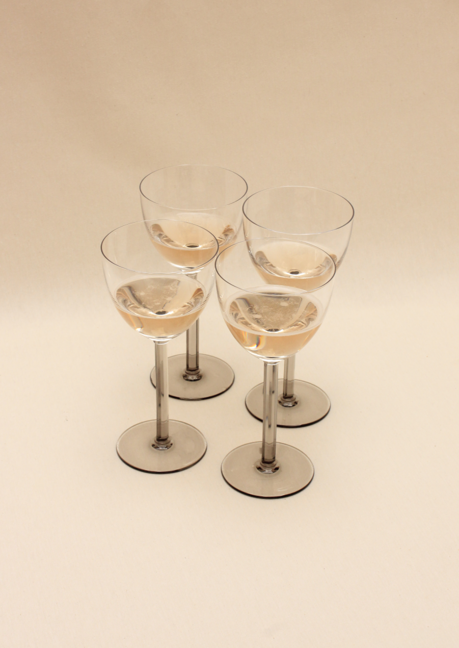 Four Boga Avante Shop Crystal Wine Glasses, handcrafted and partially filled with a light pink liquid, are elegantly arranged on a neutral beige surface in a symmetrical display that showcases contemporary design.