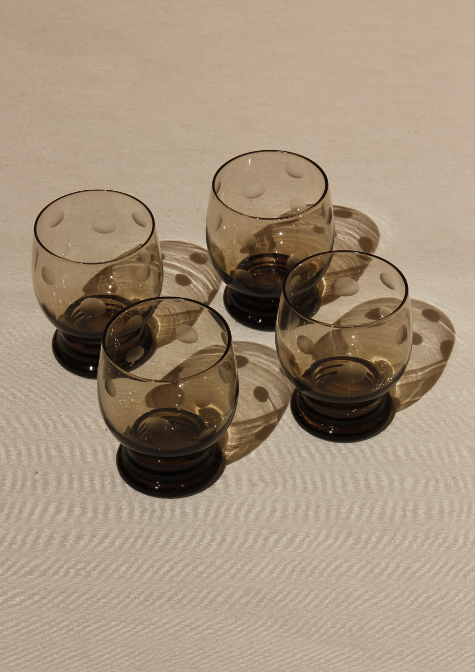 Four round, smoked glass tumblers from the Boga Avante Shop's Set of Polka Dot Glasses are arranged on a beige surface, casting soft shadows in natural light.