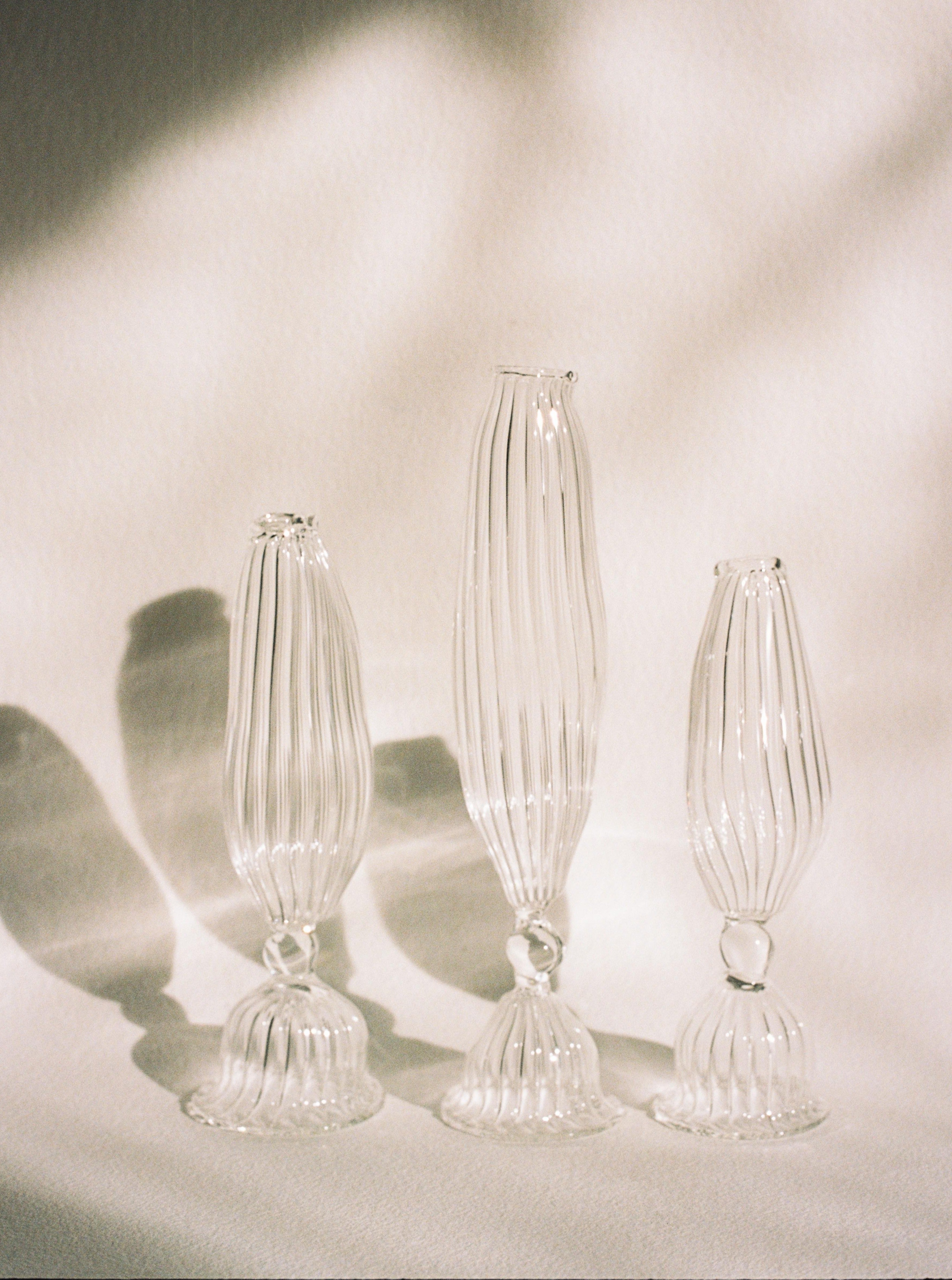 Four elegant mouth-blown glass Three Sisters Vases by Justine Menard with vertical ribbed patterns cast soft, intricate shadows on a draped cloth surface, illuminated by natural light.