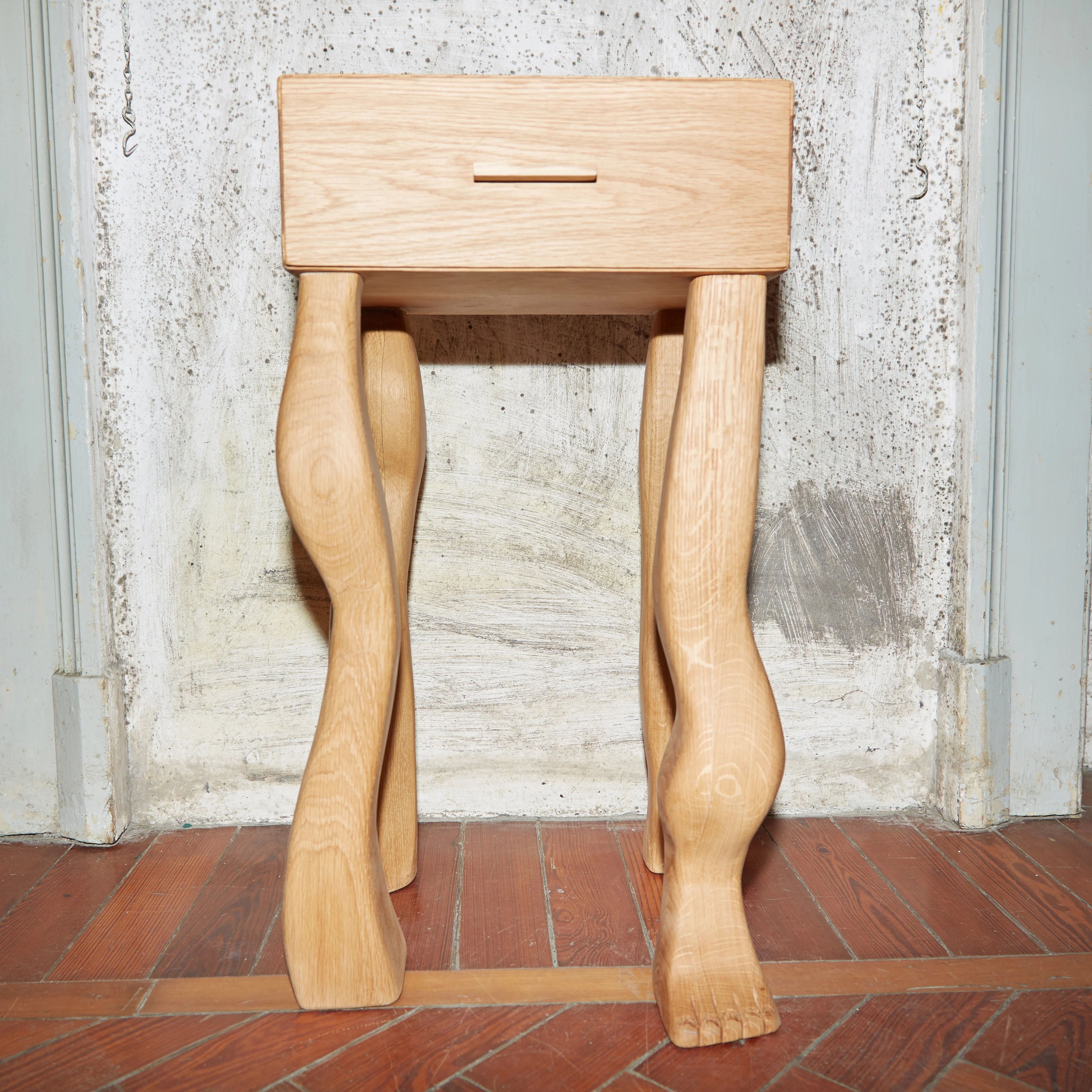 A handcrafted oak wood Foot Side Table With Drawer from Project 213A with intricately carved legs shaped like realistic human legs, standing on a tile floor against a worn wooden door.