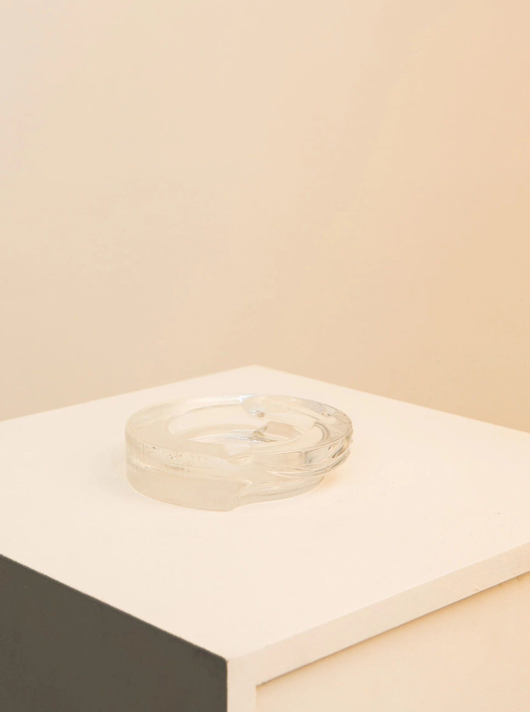 A clear glass ashtray from the Azteca Collection by Fabio Frontini for Arnolfo di Cambio 60's is placed on a white platform against a cream-colored background. The ashtray is empty and showcases a simple design. Made by Treaptyque.