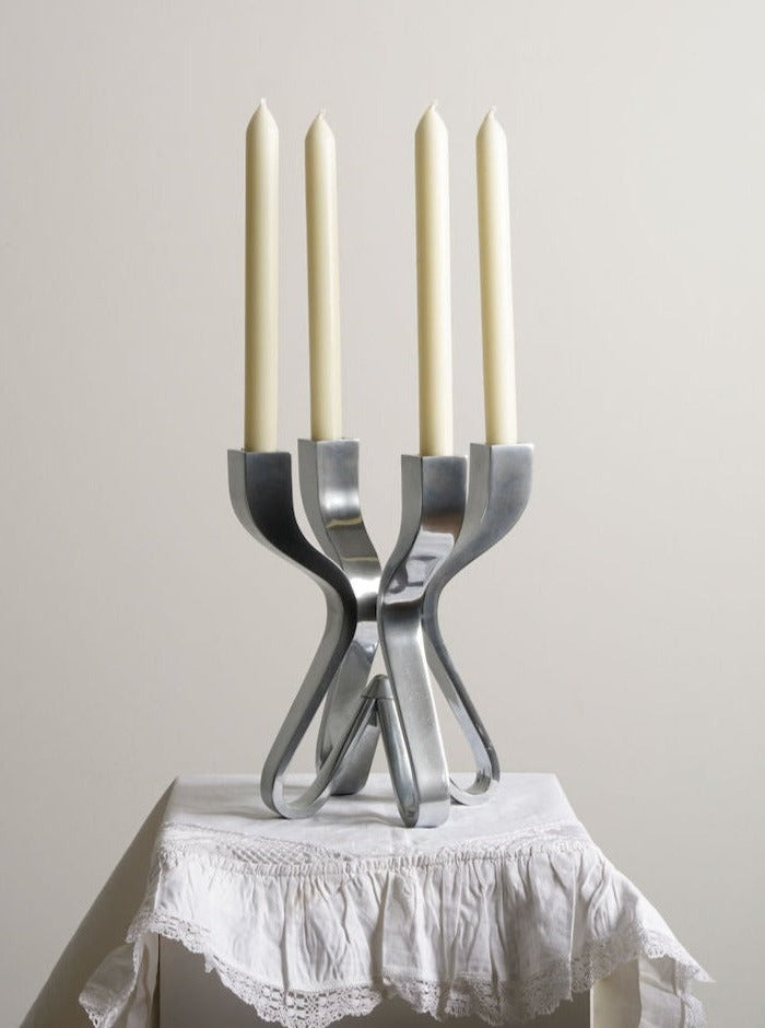 A modern, metallic candle holder by Les Objets Amsterdam, featuring four tall, unlit white candles, sits atop a small, white-draped table. The Karim Rashid-designed Umbra Candle Holder boasts a sleek, intertwined design that adds a touch of elegance to the minimalist setting.