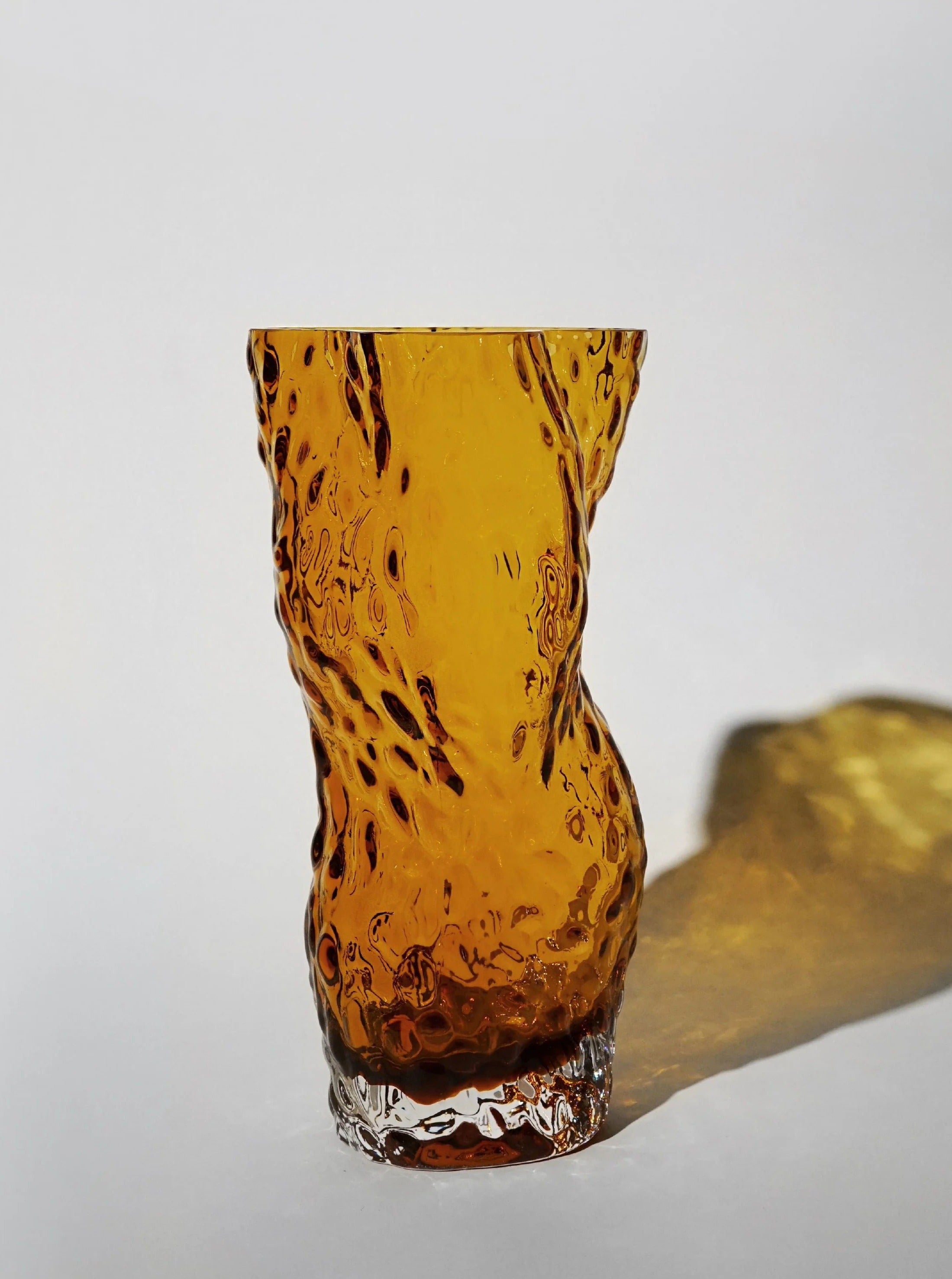 The Ostrea Rock Glass Vase in amber by Hein Studio, with its handcrafted wavy design, stands on a light surface casting a soft shadow to the right, adding sculptural elegance and striking contrast against the neutral background.