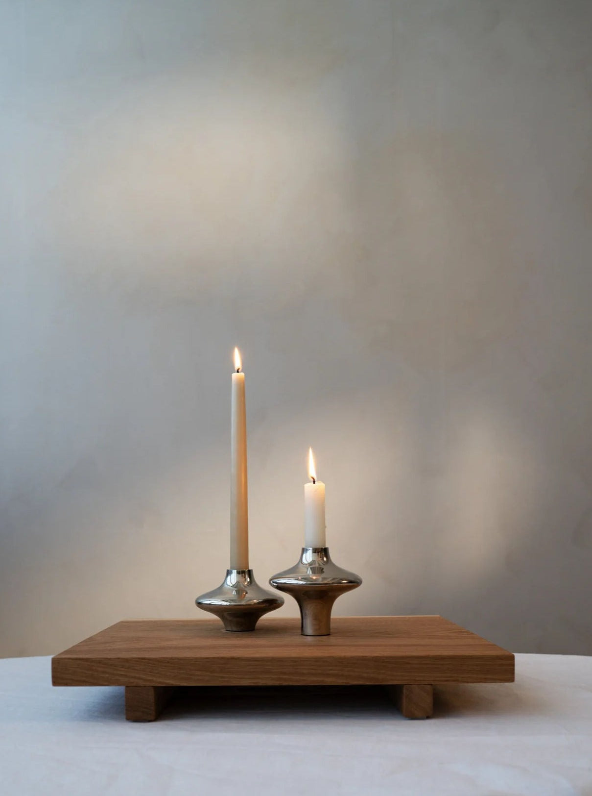 Two lit candles in sleek silver Hein Studio Doublet Candleholders standing on a wooden tray against a soft grey textured background.