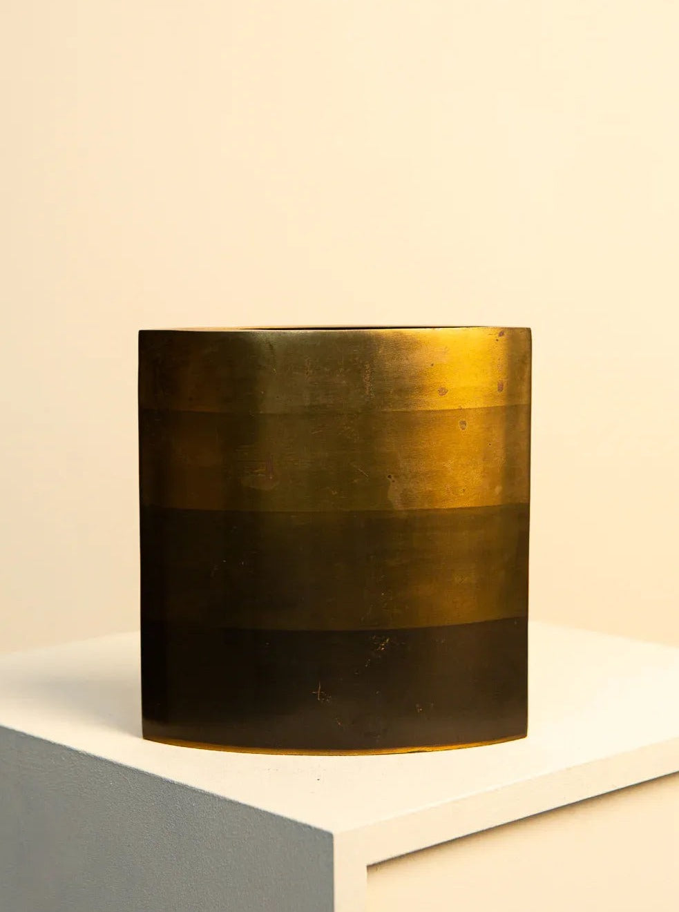A cylindrical brass and bronze vase by Treaptyque, resembling a Michael Aram vase with horizontal stripes, displayed on a white pedestal against a cream-colored background. The surface shows signs of patina and wear.