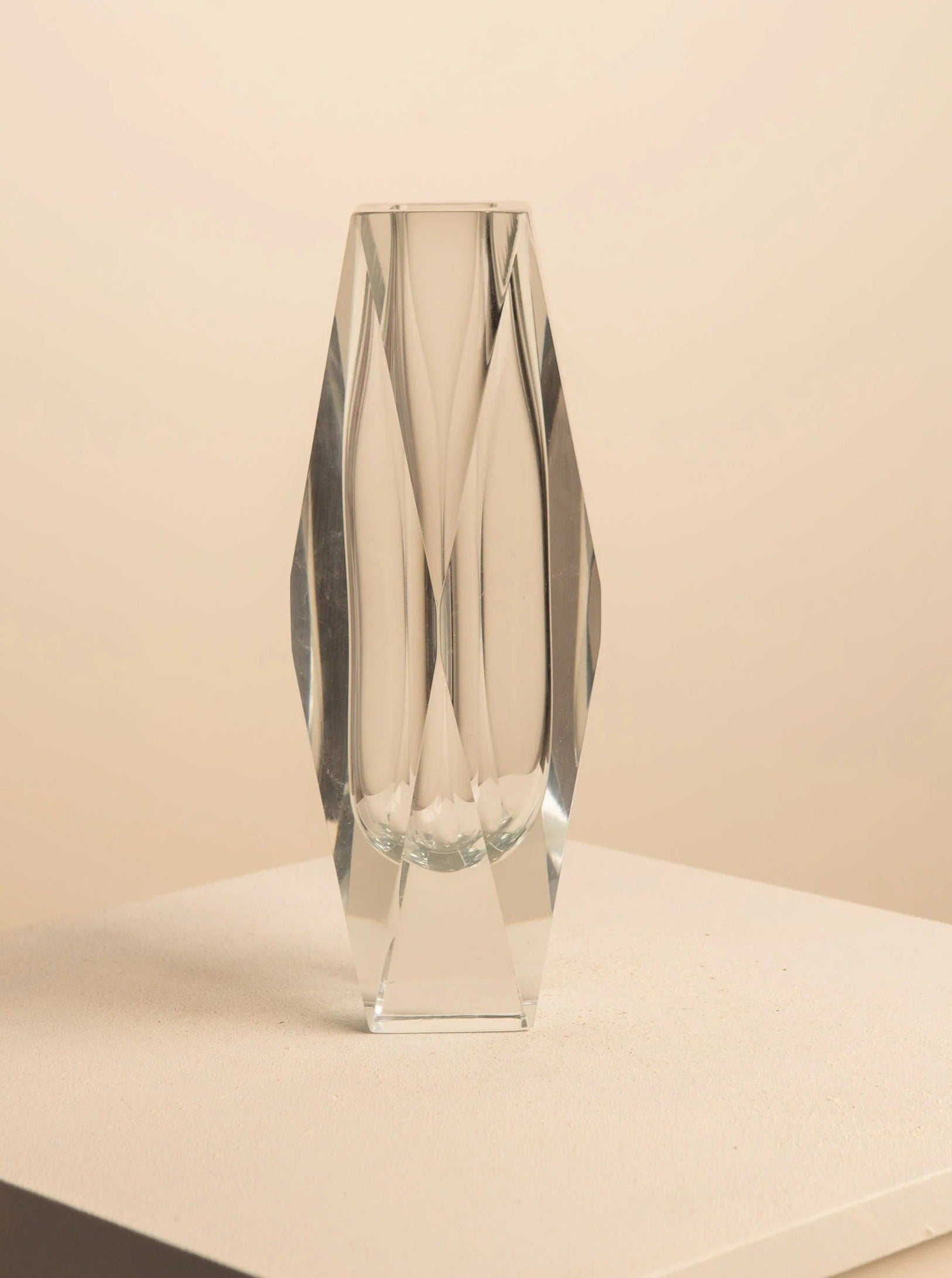 The Transparent Vase by Flavio Poli for Seguso, from Treaptyque, stands on a textured white surface and a plain beige background. Its angular, handcrafted glass design catches light beautifully, creating subtle patterns that enhance its contemporary minimalist aesthetic.