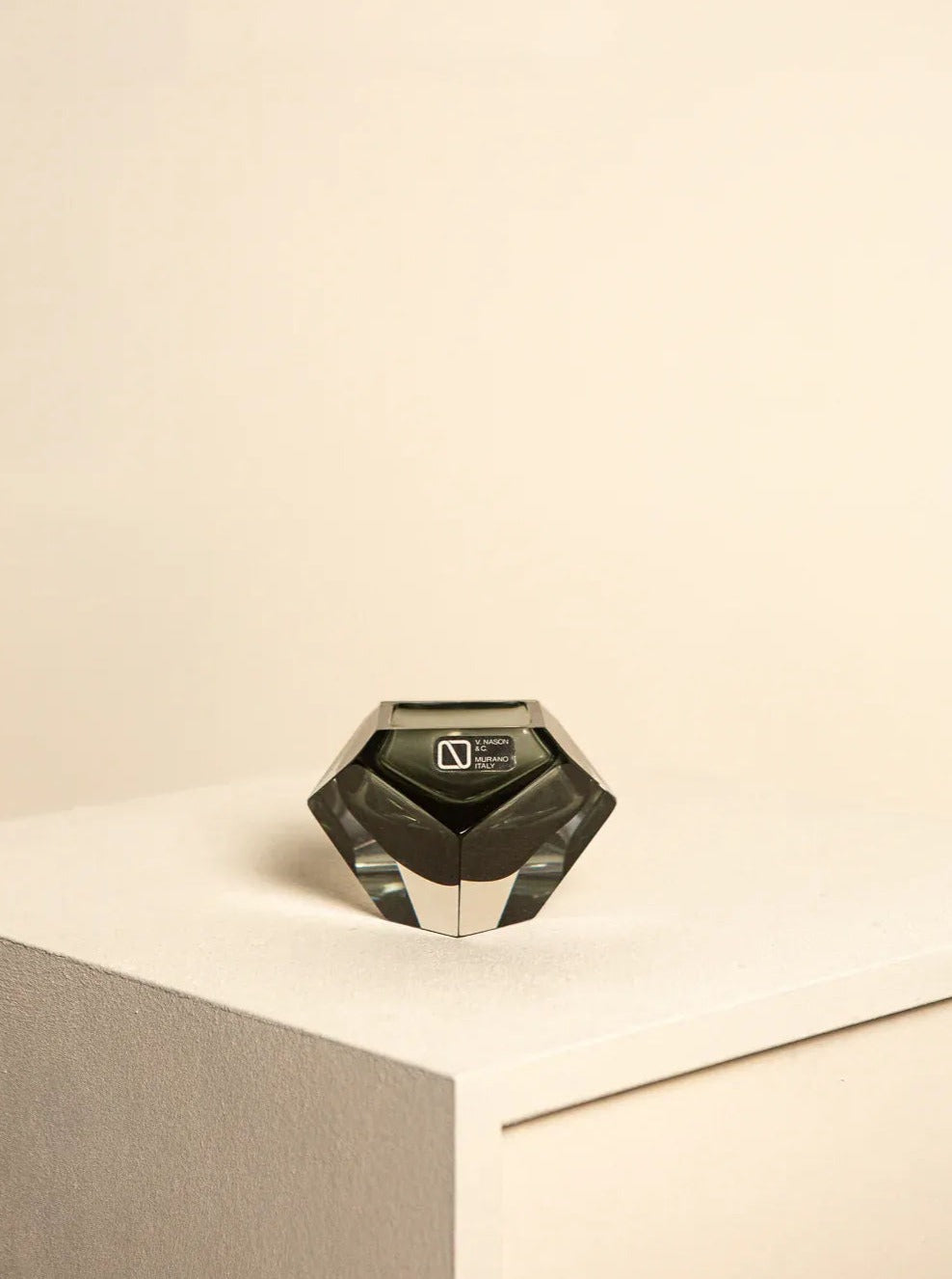 A small, black "Diamant" Vide Poche jar by Flavio Poli for Seguso from the 60's, showcasing Treaptyque's distinctive geometric design with a silver label, rests on the corner of a white minimalist table against a plain beige background.