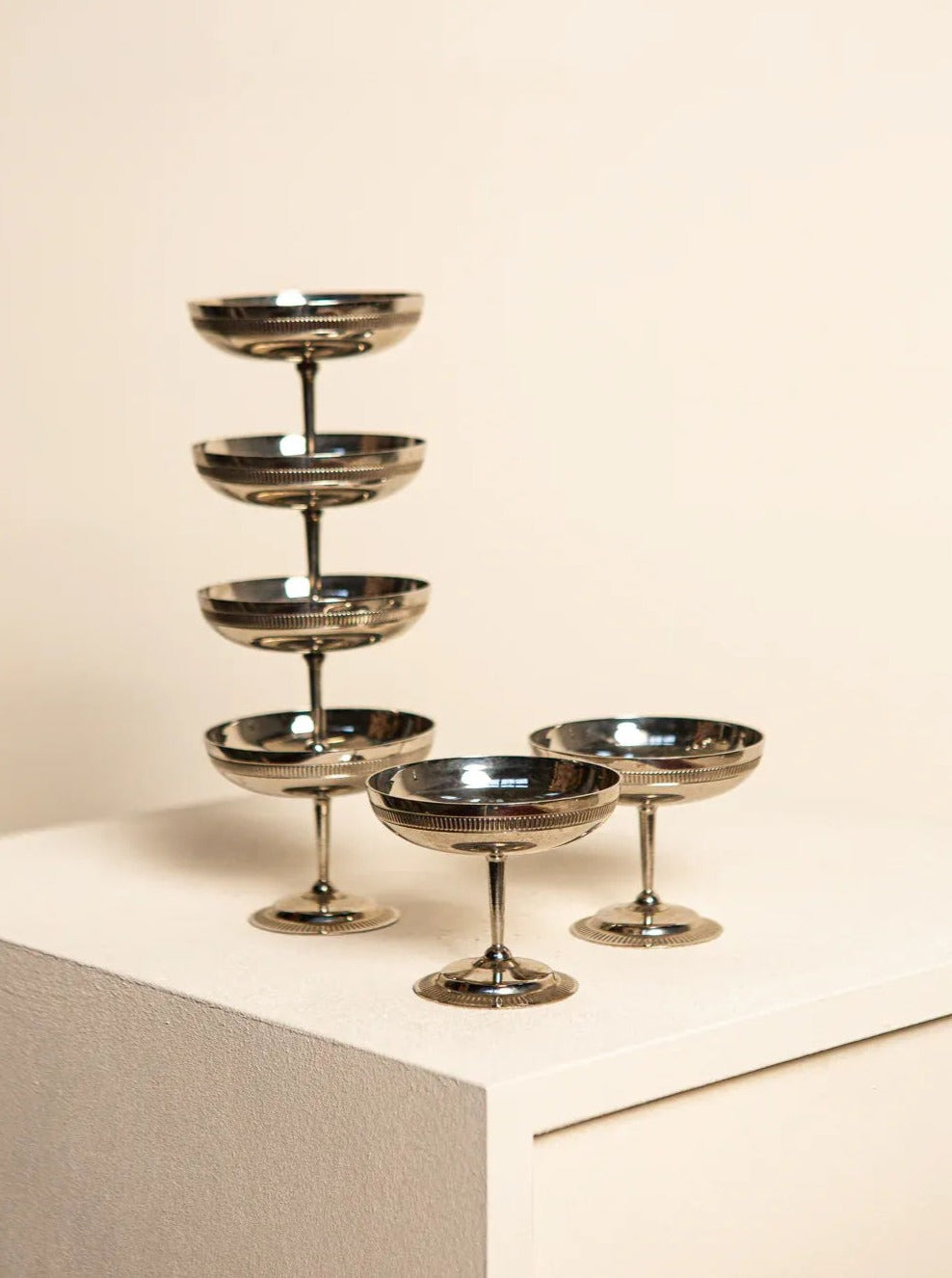 A set of elegant, Treaptyque metallic champagne coupes arranged aesthetically on a cream-colored surface, exuding retro elegance with a tower of stacked coupes in the background.